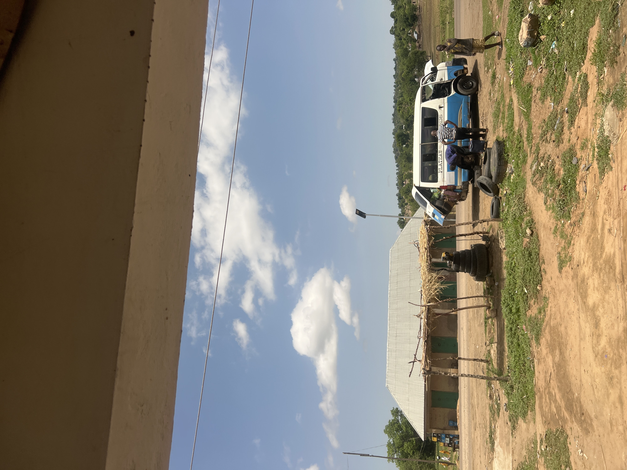 The scene is set in Sabon Layi Jessu, Nigeria, during daytime under a clear blue sky with scattered white cumulus clouds.
In the foreground, the ground is predominantly reddish-brown dirt with sparse patches of green grass and scattered litter, including plastic debris.
In the midground, to the left, stands a single-story building with a light-colored wall and a silver corrugated metal roof, featuring at least three prominent green doors or shutters. Adjacent and closer to the viewer is a rudimentary open-sided shelter constructed from several wooden poles supporting a thatched roof made of dried straw. Beneath this shelter, a stack of approximately five vehicle tires is visible. A solar-powered street light pole stands to the right of this building.
To the right in the midground, a white and blue passenger van is parked, facing slightly left. The rear hatch of the van is open, and the front hood appears to be ajar. The side of the van displays "PLATEAU EDERS" in partially obscured lettering. Three males are in the vicinity of the van: one male, dressed in a dark blue shirt and dark trousers, is crouched near the rear driver's side wheel, engaged in what appears to be a tire or vehicle repair. A second male, wearing a striped short-sleeved shirt and dark trousers, stands with his back partially to the camera, observing the first male or the vehicle. A third male, wearing a light-colored patterned shirt and shorts, stands to the right of the van, looking towards the viewer, with his right hand near his mouth. Several loose, worn tires are scattered on the ground around the van.
In the background, a paved road stretches horizontally behind the van. Beyond the road, the landscape consists of abundant green trees and distant, tree-covered hills. A utility pole with overhead wires is visible on the far left. The perspective suggests the photograph was taken from under an overhang or ceiling.