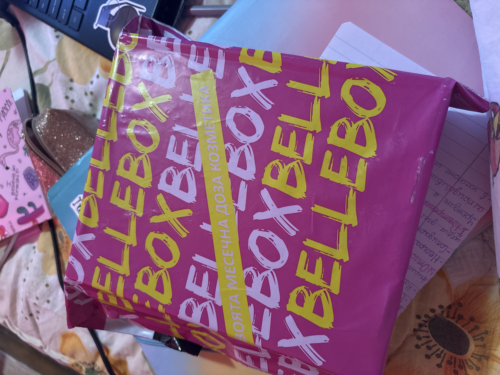 A close-up shot shows a pink package with yellow and white lettering, predominantly displaying the word "BELLEBOX" repeated in a stacked pattern. A yellow strip vertically crosses the package with Bulgarian text. The package sits on a surface with a floral patterned sheet. Adjacent to the package, a notebook with handwritten notes in Bulgarian is visible. Other items like a keyboard, a glittery pouch, and makeup palettes are partially visible in the background. The image does not reveal any information that indicates the location in the city of Garmen, Bulgaria.