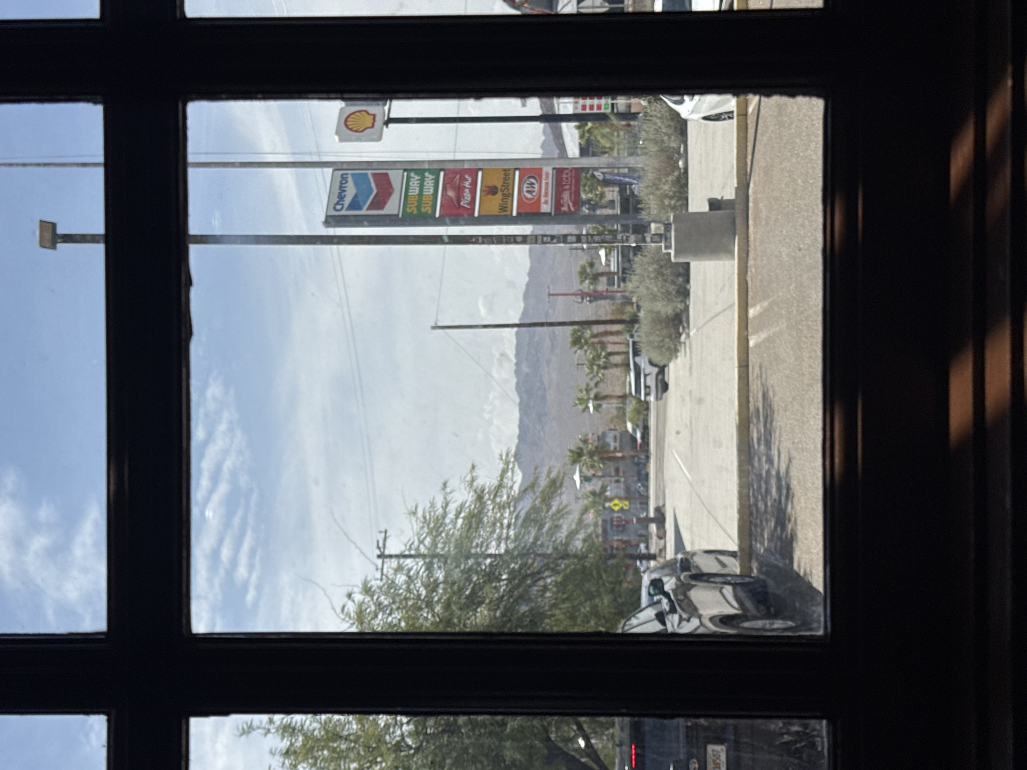 The image shows a view through a window frame, looking out onto a street scene in Baker, California, United States. The foreground is dominated by the dark, wooden window frame. The middle ground features a road with several vehicles, including a black SUV and a white car. There are also palm trees, desert scrub, and a concrete bollard. In the background, a tall signpost displays logos for Chevron, Subway, Pizza Hut, WingStreet, and A&W. A Shell gas station sign is also visible. Mountains are visible in the far distance, and the sky is partly cloudy. The scene appears to be a typical roadside view in a desert town.