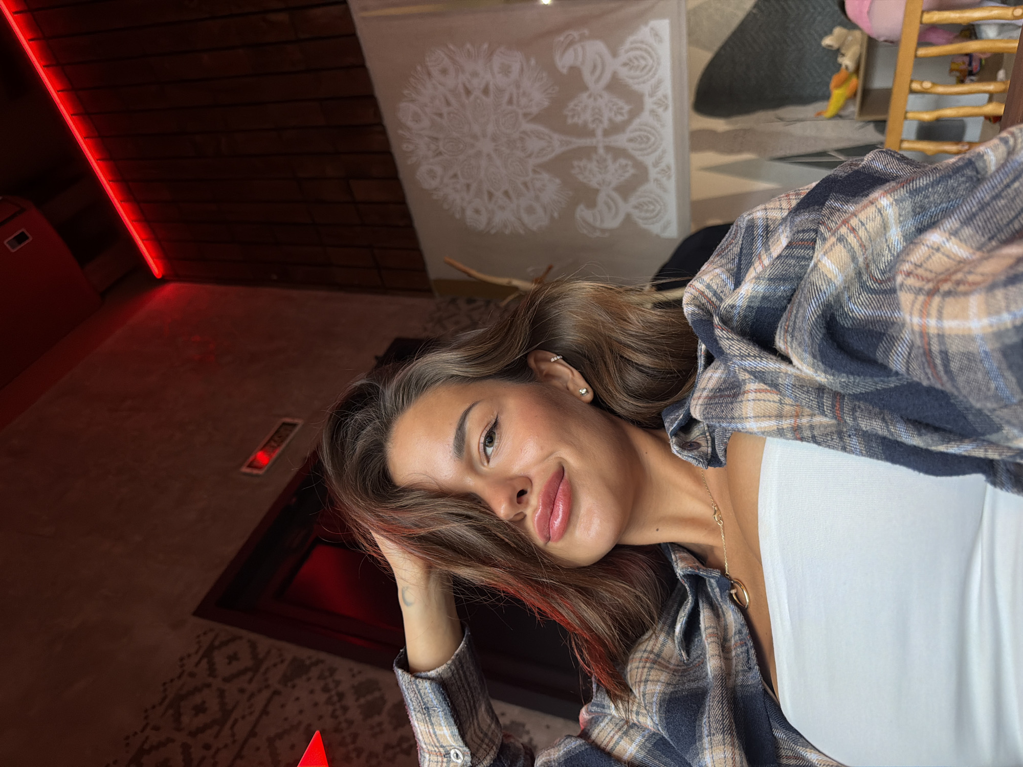 A young woman with long, wavy brown hair is taking a selfie in what appears to be an indoor setting. She is wearing a white strapless top and an open plaid shirt. Her left hand is resting on her head, and she is looking at the camera with a slight smile. Her makeup is noticeable, with defined eyeliner and prominent lip gloss. In the background, there is a red-lit wall with a patterned texture and a wooden paneling with a red LED strip light. A piece of fabric with a white floral or tree-like design is hanging on the wall to the right. The overall lighting is dim and moody, with red hues dominating the scene. It is impossible to determine the specific location within Krasnoyarsk, Russia from the provided image.