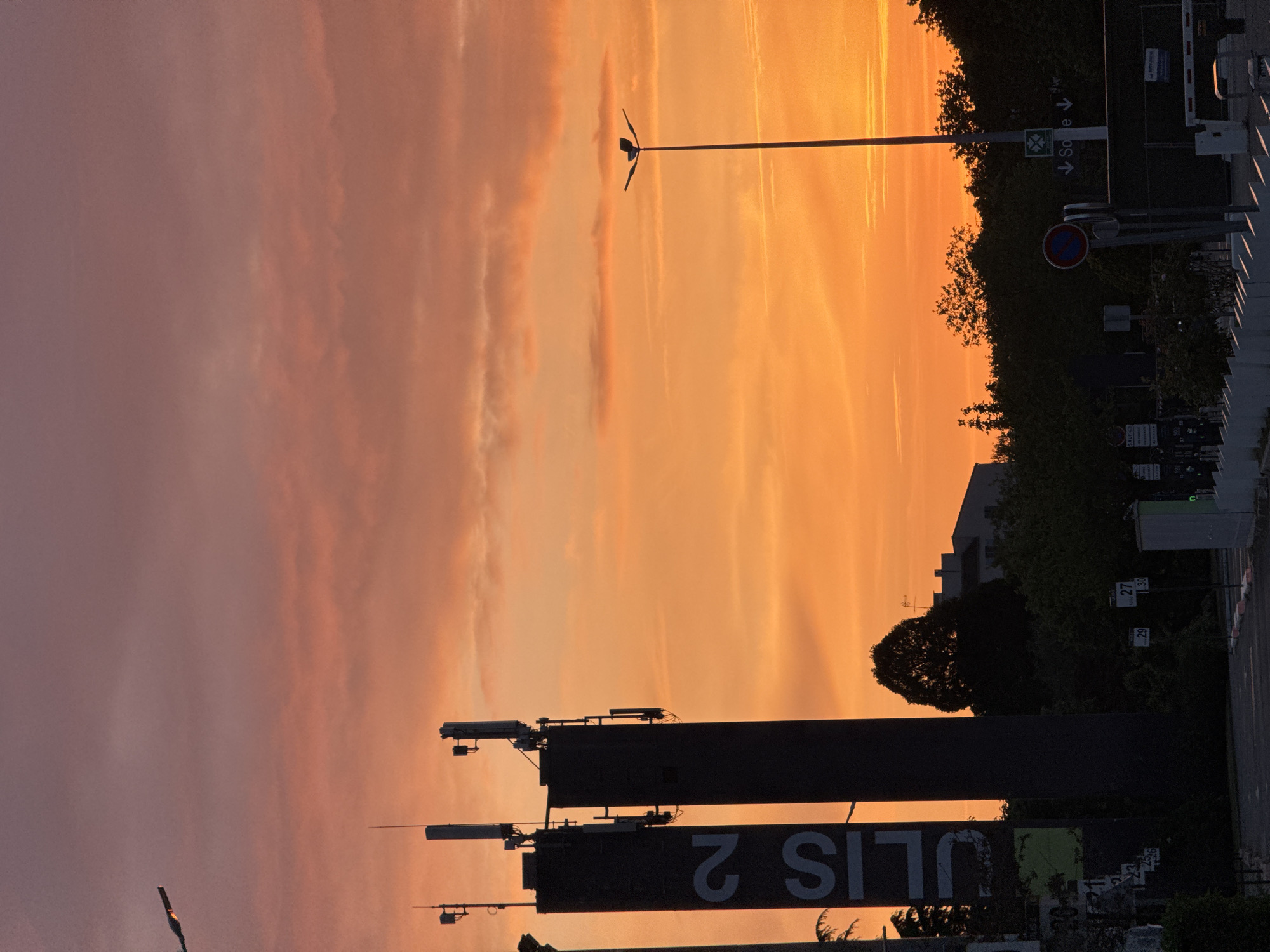 The image is a sideways view of the city of Saint-Jean-de-Beauregard, France, at sunset. The sky is a mix of pink and orange. In the foreground are the silhouetted objects of the city, including buildings, signs, and a lamp post. The sign reads "ULIS 2." There is foliage. The objects in the city fade into the background due to the low light.