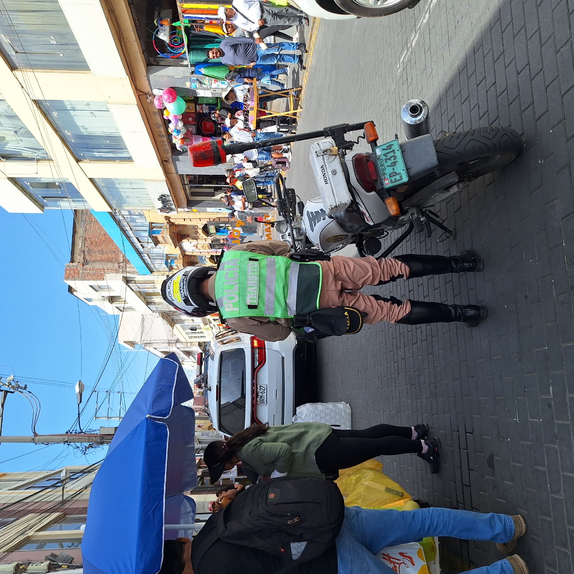 The image captures a bustling street scene, likely in a commercial area of Peru, on a bright, sunny day. The primary subject is a police officer wearing a light brown uniform with black boots, a green reflective vest, and a helmet. The vest clearly displays "POLICÍA TRÁNSITO", and the helmet also shows "TRANSITO", "POLICÍA", "AN", "AF", and "LAN". The officer is standing beside a white Honda motorcycle with a Peruvian license plate "EP-4314".
In the foreground, another person is partially visible, wearing blue jeans and light brown shoes. Closer to the viewer, a person in a green shirt and black cap is visible, possibly sitting or crouching, near a white van with a partially visible license plate "BRM-627". A blue market umbrella is also prominent in the foreground.
The background reveals a lively street lined with multi-story buildings, some with large windows. Several people are visible, appearing as pedestrians or vendors, some standing around market stalls displaying various goods like colorful balloons and other items. Overhead, numerous power lines crisscross against a clear blue sky. The street is paved with dark grey brick-like tiles, and strong shadows indicate midday sun. The scene suggests a busy urban environment with active commerce and daily life.