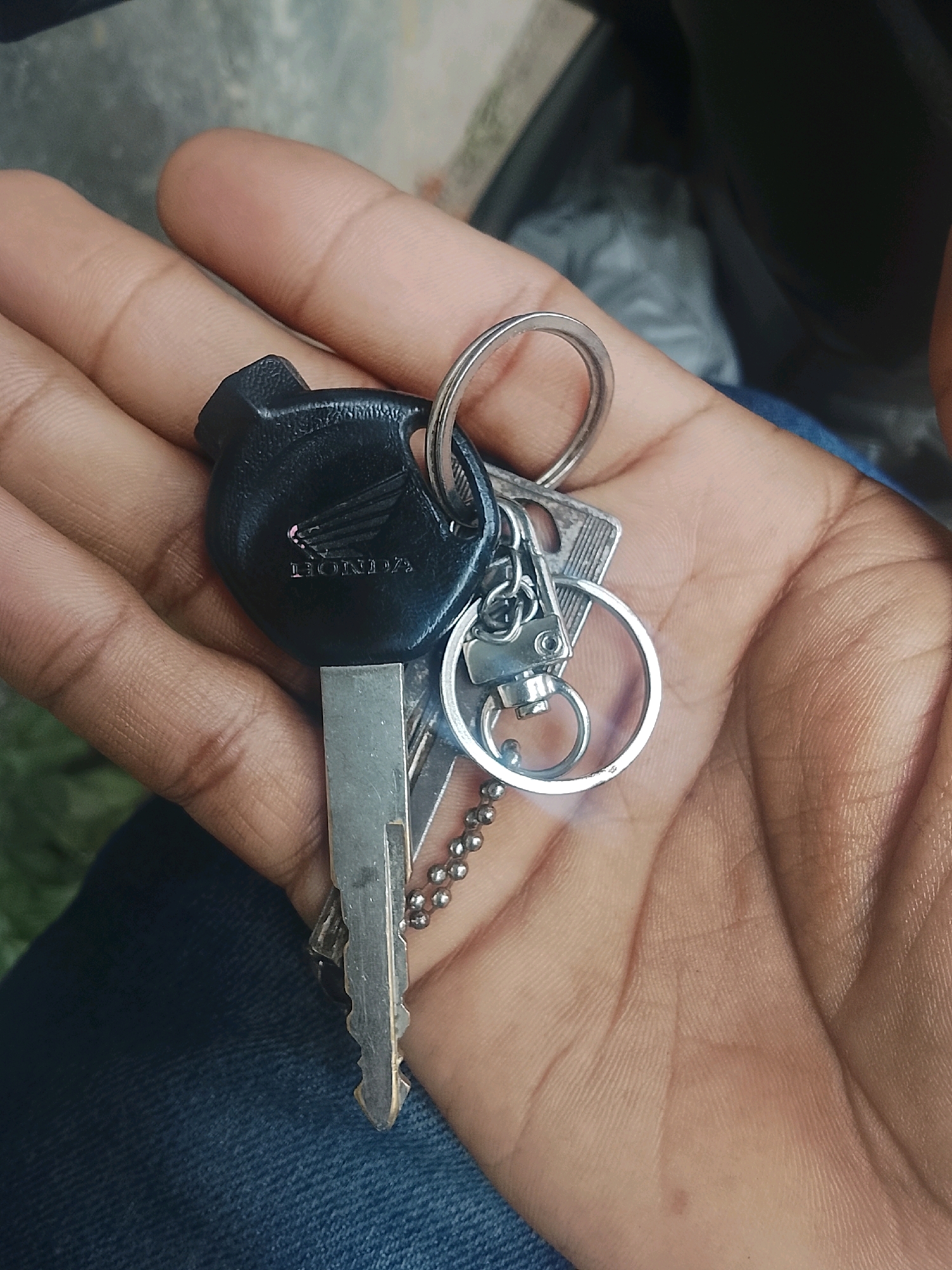 In Kertahayu, Indonesia, a close-up shot captures a hand holding a set of keys. The hand, with its visible lines and texture, is the central focus against a blurred background that suggests an outdoor setting with muted colors. The keys are attached to a black fob featuring the "HONDA" logo, indicating they are likely for a Honda motorcycle. A silver ring and a beaded chain connect the fob to at least two metal keys. One key is silver and shows signs of wear, with a distinct pointed tip. The other key is partially obscured but appears similar in style. The lighting is somewhat diffused, casting gentle shadows that highlight the details of the hand and the metallic sheen of the keys. The overall impression is one of everyday utility, a common scene in any locale, including Kertahayu.