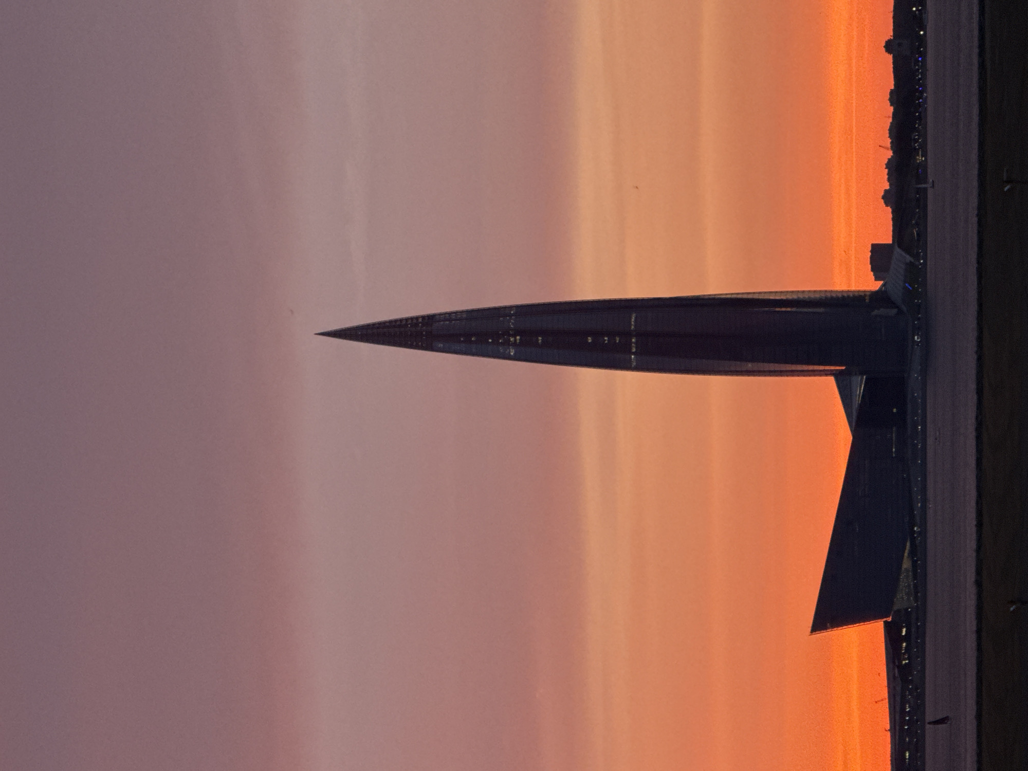 The image depicts the Lakhta Center skyscraper in Saint Petersburg, Russia, silhouetted against a vibrant sunset. The sky transitions from a deep purple at the top to bright orange and pink hues near the horizon. The modern, needle-like skyscraper dominates the center of the frame, its glass facade reflecting the colors of the sky. To the left of the skyscraper, a modern, angular building is visible, also silhouetted. In the foreground, a dark, flat expanse, likely a body of water or land, separates the viewer from the structures. The overall scene is one of an urban landscape at dusk, with the striking architecture of the Lakhta Center being the primary focal point.
