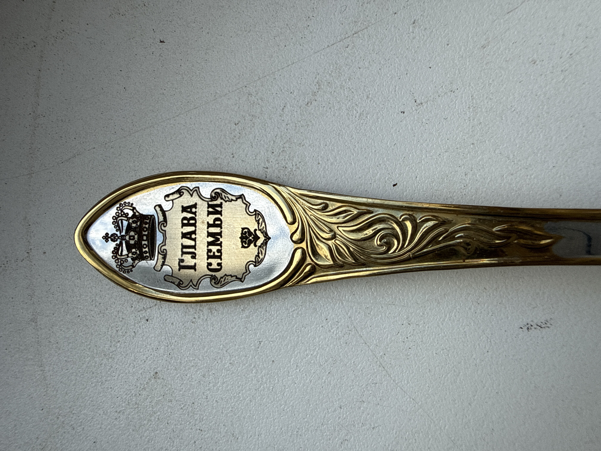 A decorative spoon is placed on a textured, light-colored surface. The spoon features an ornate design at the top, including a crown and intricate lettering in what appears to be Cyrillic. Below the crown and lettering, there is a detailed floral engraving that runs down the length of the spoon's handle. The spoon is primarily gold in color with some silver accents. The lighting suggests a bright environment, casting subtle shadows. The background is out of focus, emphasizing the spoon as the central subject. The location is not discernible from the image.