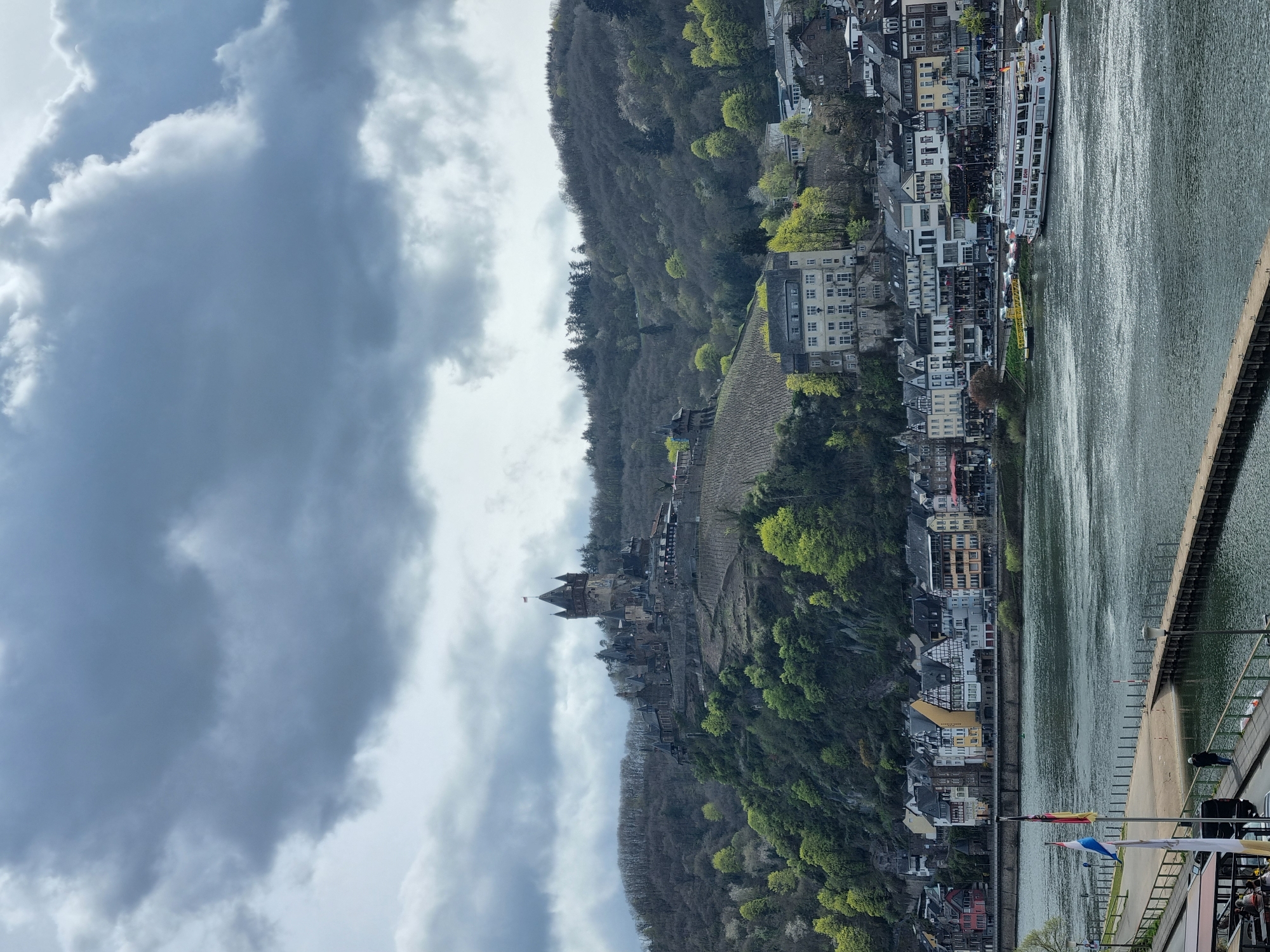 The image shows Cochem, Germany, on a cloudy day. The Reichsburg Cochem castle is perched atop a hillside covered in vineyards and trees. A river flows along the base of the hill, lined with buildings of the town. A dock or pier runs along the river's edge. A large passenger boat is docked along the river.