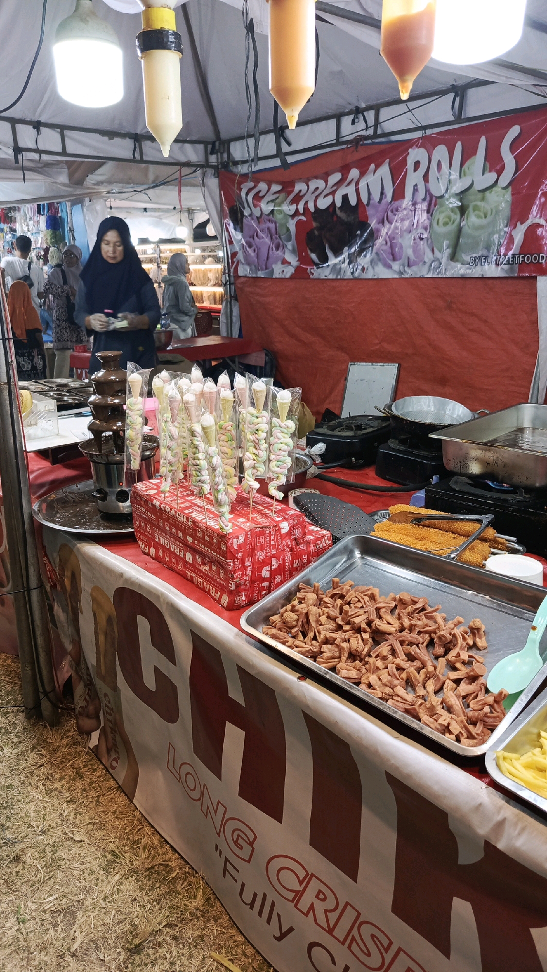 The image depicts a street food stall in Tegal, Indonesia, specializing in ice cream rolls. The stall is set up outdoors, likely at a market or event, with a red tent covering the area. A large red banner with the words "ICE CREAM ROLLS" is prominently displayed at the back, featuring colorful illustrations of rolled ice cream.
In the foreground, a vendor wearing a black hijab is seen preparing food. In front of her, a chocolate fountain is active, with a cascade of chocolate flowing down. To the right of the fountain, several skewers of colorful marshmallows topped with ice cream cones are arranged on a red display box. A metal tray filled with small, fried, brown food items, possibly sausages or similar snacks, is placed on the counter. Another tray containing yellow, thinly sliced food, likely fries or another side dish, is partially visible to the right.
The stall is decorated with a large banner that reads "CHIC LONG CRISPY" with the slogan "Fully Crispy" underneath. The ground appears to be covered with dry grass and straw. In the background, other stalls and people can be seen, suggesting a bustling market environment. The overall scene captures a lively outdoor food vending operation.