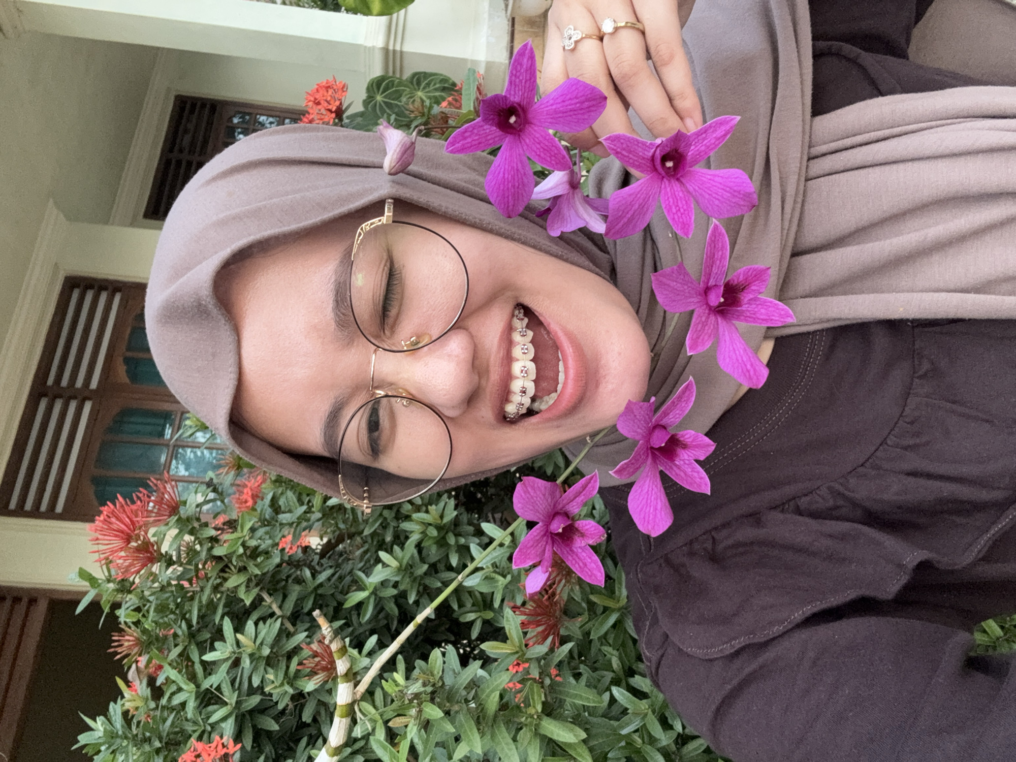A young woman wearing a beige hijab and round glasses is taking a selfie outdoors. She has braces on her teeth and is winking, holding pink orchids in her right hand. Behind her is lush green foliage with red flowers. The background also shows a building with wooden window frames and a porch. She is wearing a dark brown shirt. The scene appears to be in the city of Gadingrejo, Indonesia.