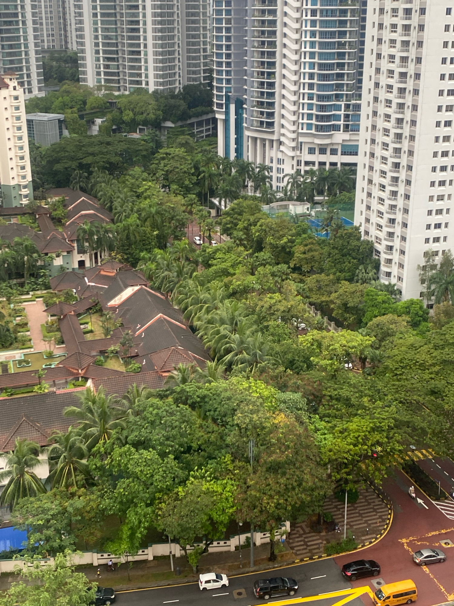 The image depicts a high-angle view of a dense urban landscape in Kuala Lumpur, Malaysia, characterized by a juxtaposition of high-rise residential buildings and lower-rise, tiled-roof structures surrounded by lush greenery. Dominating the background are numerous modern, tall apartment buildings with white facades and repetitive window patterns. In the midground and foreground, a complex of low-rise buildings with dark, sloped tiled roofs and some exposed wooden beams are visible, suggesting a residential or communal complex. Extensive tree cover, including palm trees, fills the spaces between these structures, creating a sense of a green city.
Several vehicles are present on the paved roads visible in the lower portion of the image. These include a white sedan, a black SUV, a black sedan, a silver sedan, and a yellow school bus. A few individuals are also discernible on a sidewalk or path near the low-rise buildings and along the road. One person appears to be walking, while others are too small to determine their actions definitively. The environment appears to be a developed urban area with well-maintained roads, sidewalks, and landscaping, suggesting a residential or commercial district within Kuala Lumpur. The sky is overcast, contributing to a somewhat muted color palette.