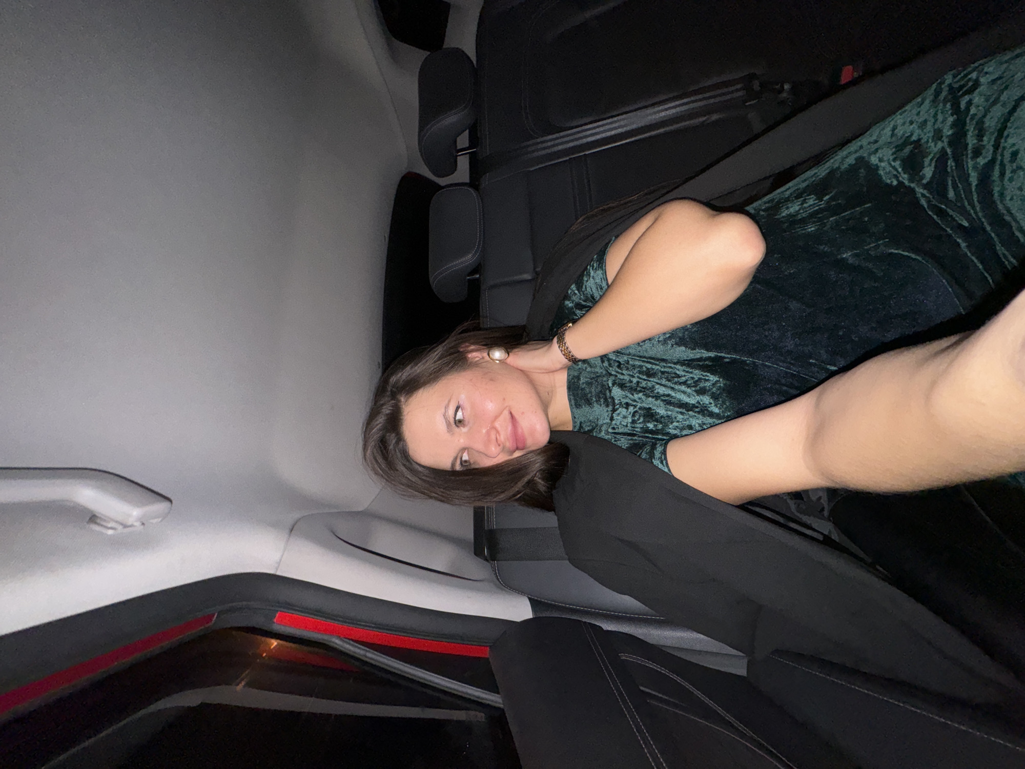 A woman wearing a green, velvet dress is sitting in the back seat of a car. She is angled towards the camera and appears to be looking at the photographer. She is wearing a gold bracelet. The interior of the car is mostly black, with some grey. There is a red trim near the car window. The location is possibly Akulovo (Gp Kubinka), Russia.