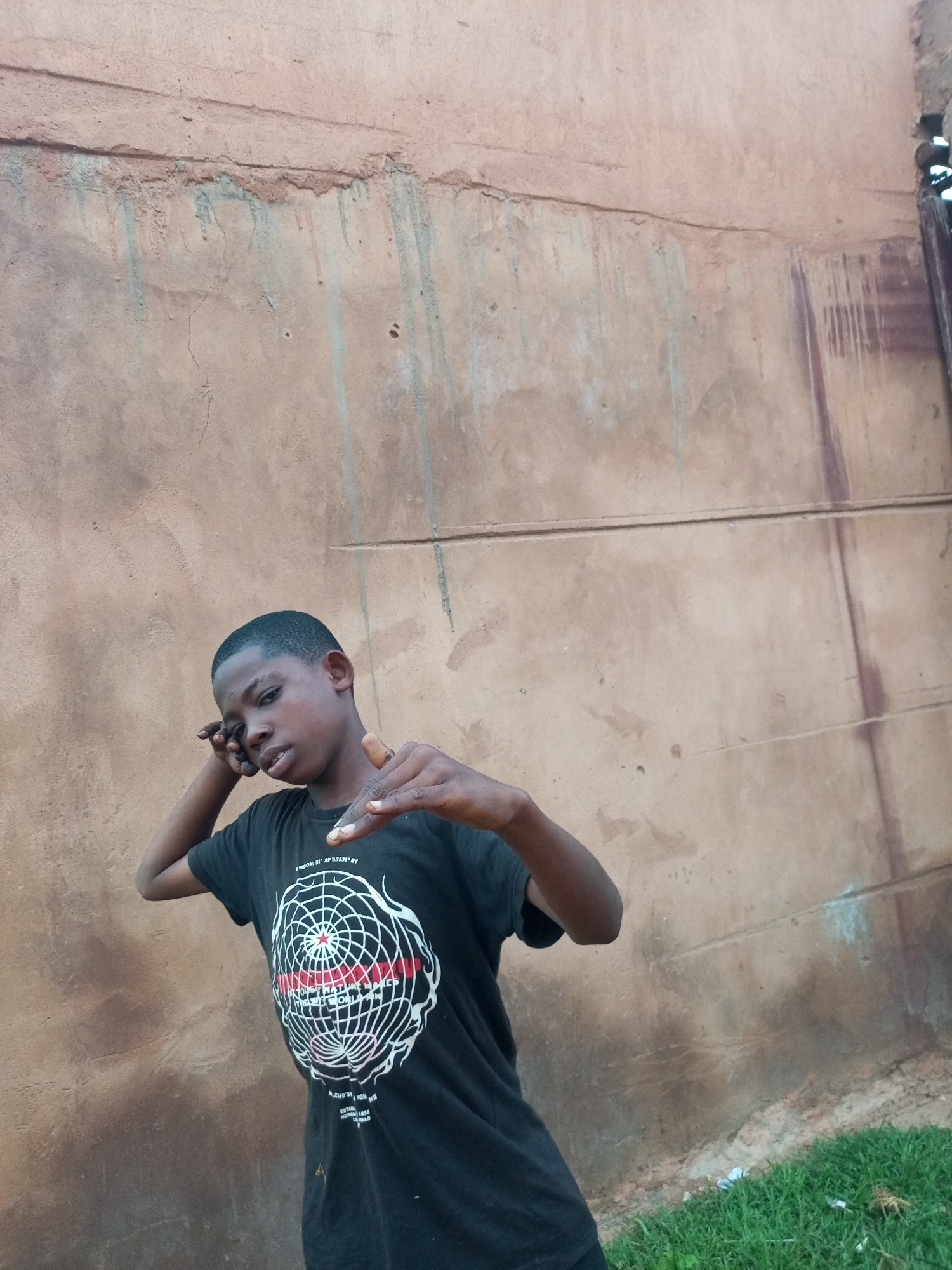 A young Black male is posing for a photo against a weathered, light brown concrete wall. The wall has streaks of green and brown discoloration and appears to be in disrepair with cracks and a rough texture. The ground in the foreground is covered in green grass with some white debris. The male is wearing a black t-shirt with a white circular design featuring lines and dots that resembles a globe or a net. He has short, dark hair and is looking directly at the camera with his right hand near his ear, as if holding a phone, and his left hand extended forward with his fingers pointing. The lighting suggests it is daytime. The location is identified as Tempure, Nigeria.