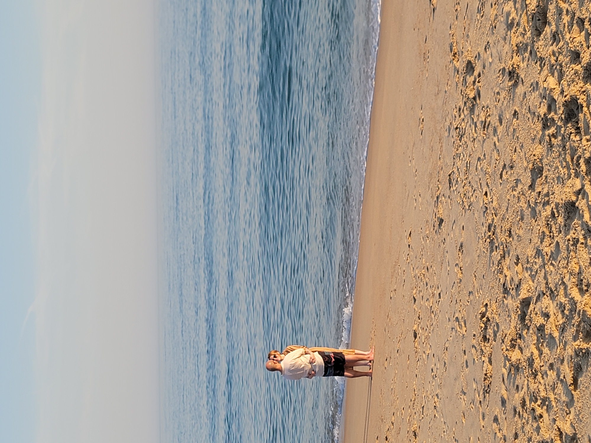 This media file depicts a serene beach scene in Quarteira, Portugal. Two individuals are standing on the wet sand near the water's edge, facing the ocean. The person in the foreground, likely a male, is bald and wearing a light-colored short-sleeved shirt and dark shorts. He is embracing another person, who appears to be a female, from behind. The second person is wearing a light-colored jacket or shawl over their clothing.
The ocean is a deep blue with gentle waves lapping at the shore, indicating calm weather. The sandy beach is textured with footprints and a few patches of dried seaweed or debris. The light suggests it is either early morning or late afternoon, with the sun casting long shadows and illuminating the sand with a warm, golden glow. The sky is mostly clear, with a hazy or foggy band along the horizon, possibly indicating the proximity of land or a change in weather.
The overall atmosphere is peaceful and intimate, suggesting a moment of quiet companionship shared by the two individuals. There is no visible text in the image.