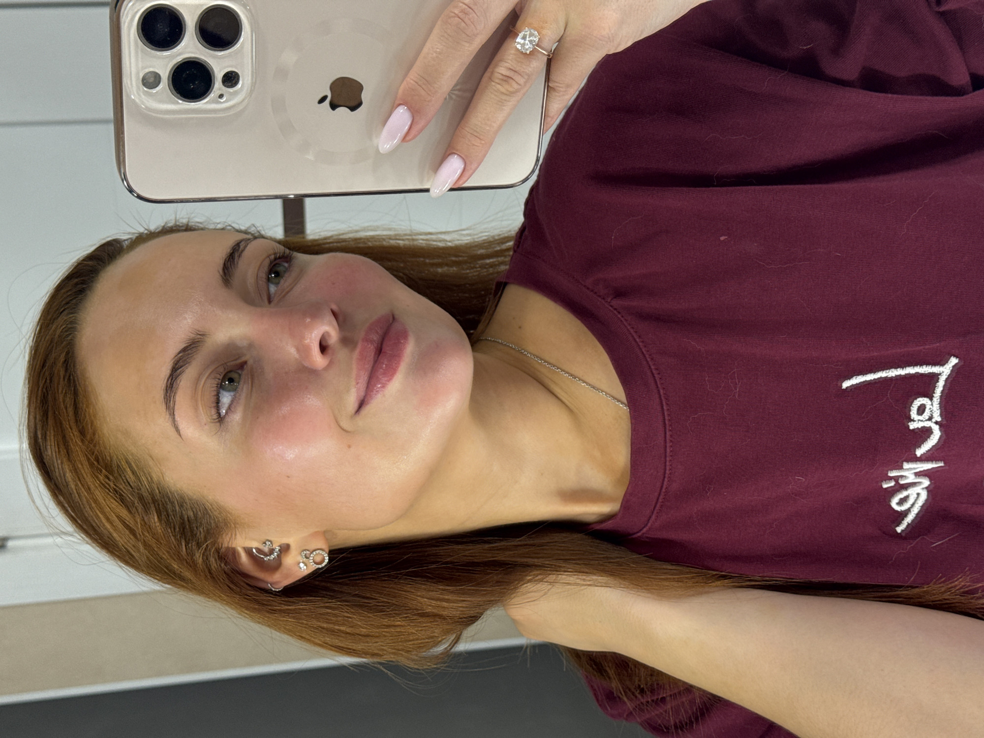 A young woman is taking a selfie with an iPhone. The phone is in a clear case, revealing the Apple logo and the triple lens camera system. The woman is wearing a maroon t-shirt and a diamond ring on her left ring finger. She has light brown hair and multiple earrings in her left ear. The location is most likely indoors, given the even lighting and the background of neutral-colored walls. There is no information provided to indicate that this image was taken in the city of Chaikovsky, Russia.