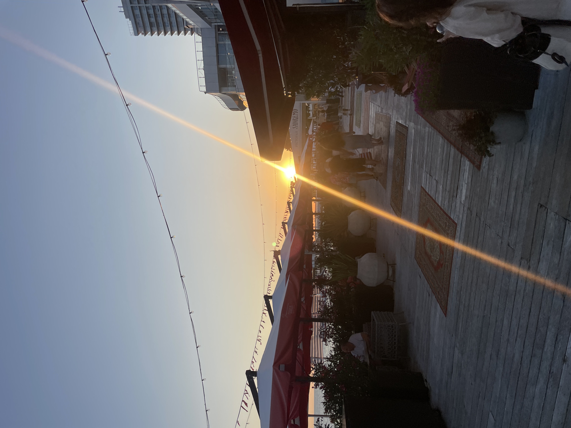 The image shows a sunset scene at a seaside cafe in Sochi, Russia. The sun is low on the horizon, casting a warm glow across the sky and the water. In the foreground, there is a wooden deck with several large, red and white striped umbrellas providing shade for outdoor seating. People are seated at tables under the umbrellas and walking along the deck. In the background, there is a modern building with multiple stories. A string of lights is visible across the top of the image, and a bright sunbeam cuts diagonally across the frame. The overall atmosphere is relaxed and inviting.