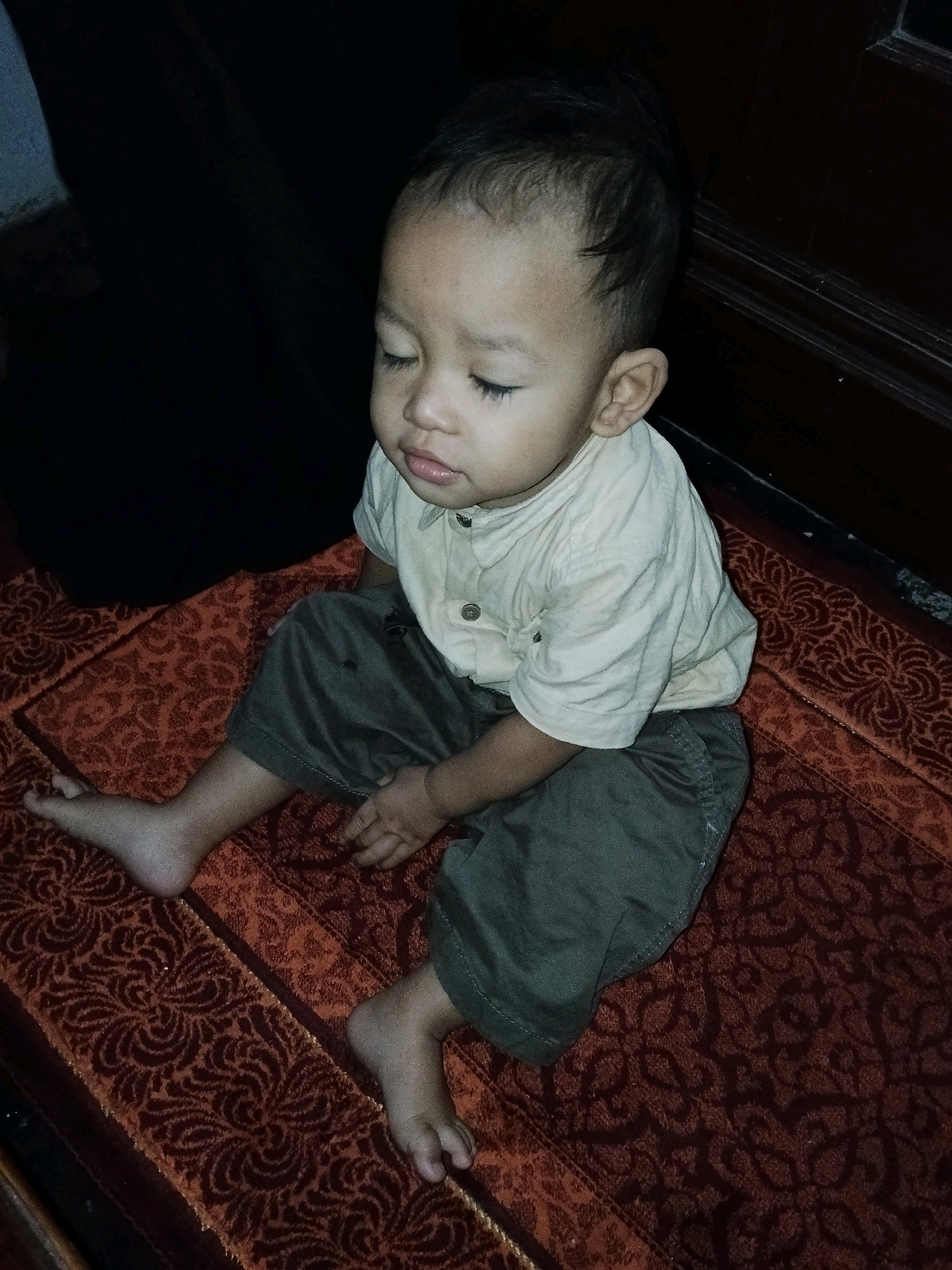 A young child with closed eyes and a light-colored collared shirt and dark pants is sitting on a red patterned rug. The child's head is tilted slightly to the left. The background is dark, with a glimpse of dark fabric on the left and a dark wooden surface on the right. The image appears to be taken indoors.