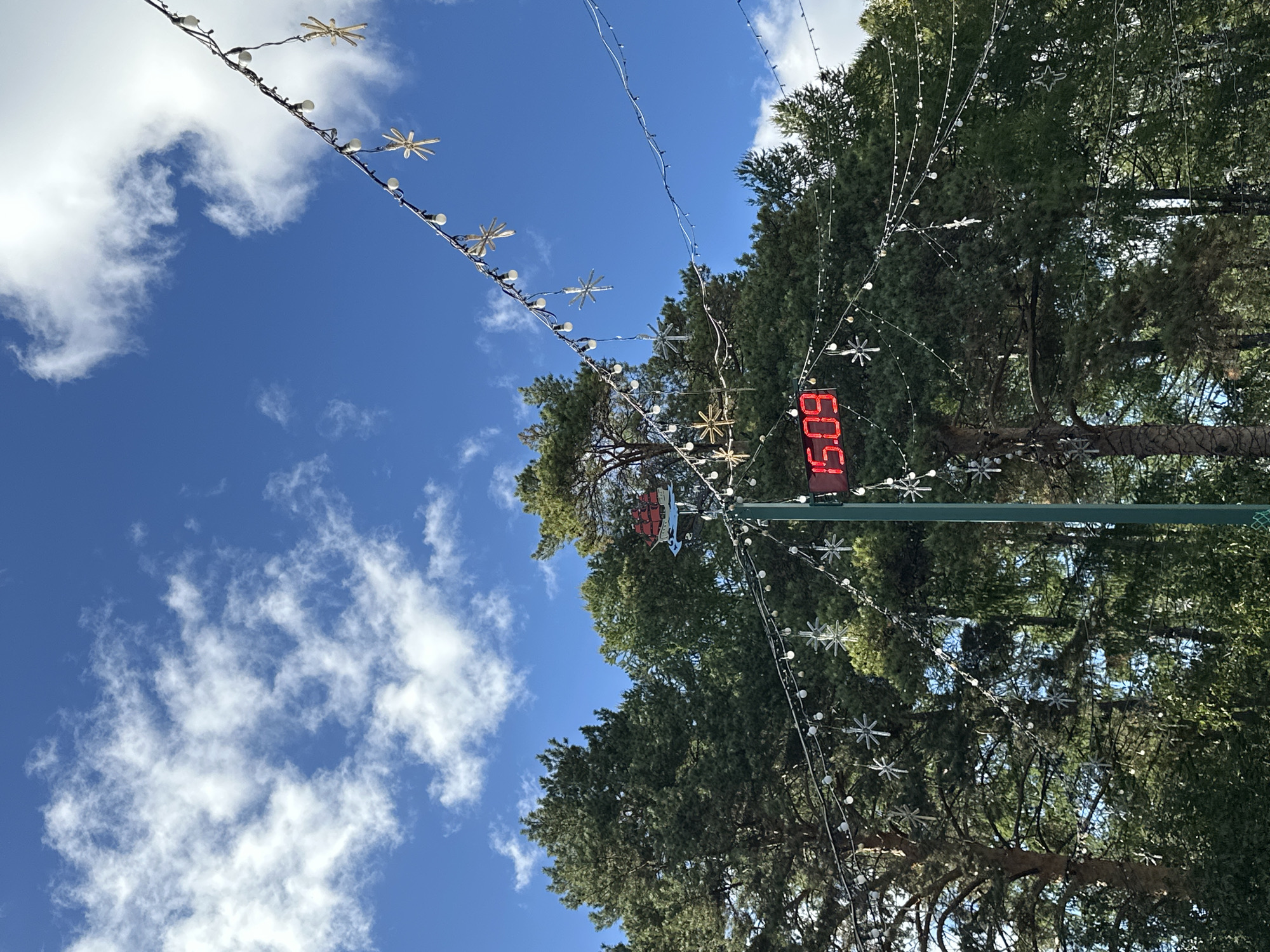 An outdoor, low-angle, medium shot captures a tall, green pole with a red digital clock displaying "15.09" and a silhouette of a ship on a blue wavy background. String lights with small white bulbs and snowflake ornaments are draped across the frame, hanging from wires strung between unseen points. The background is filled with dense evergreen trees under a bright blue sky with scattered white clouds. The scene is likely set in a park or outdoor area.