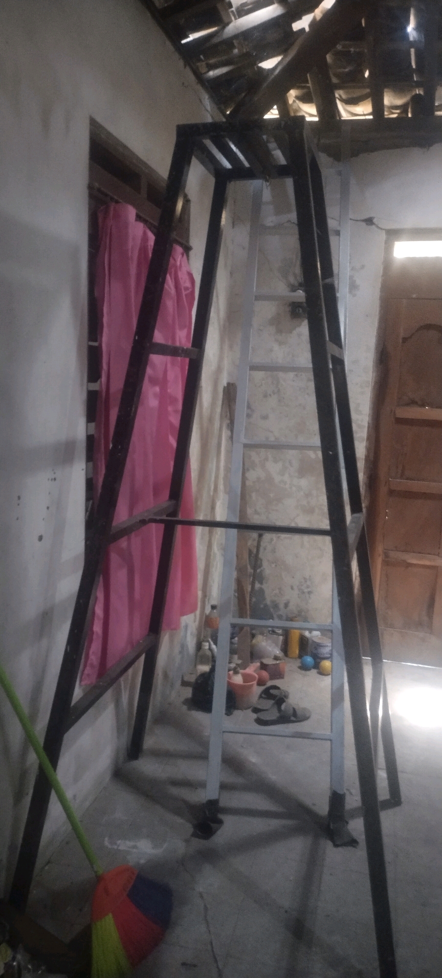 The image depicts a scene inside a room with an unfinished ceiling, exposed wooden beams, and a wall with cracked plaster. A large, A-frame metal structure, possibly a homemade ladder or scaffolding, is the central focus. It has a dark metal frame and lighter-colored rungs. To the left of the structure, a pink curtain hangs near a window. On the floor, to the right of the structure, are several objects including a red bucket, a pair of sandals, some plastic bottles, and a yellow bucket. A broom with a green handle and colorful bristles is leaning against the left side of the metal structure, near the floor. The lighting suggests an indoor environment with natural light coming from a window and possibly from an opening in the ceiling. The overall impression is one of a work in progress or a storage area. This scene is located in Daleman, Indonesia.