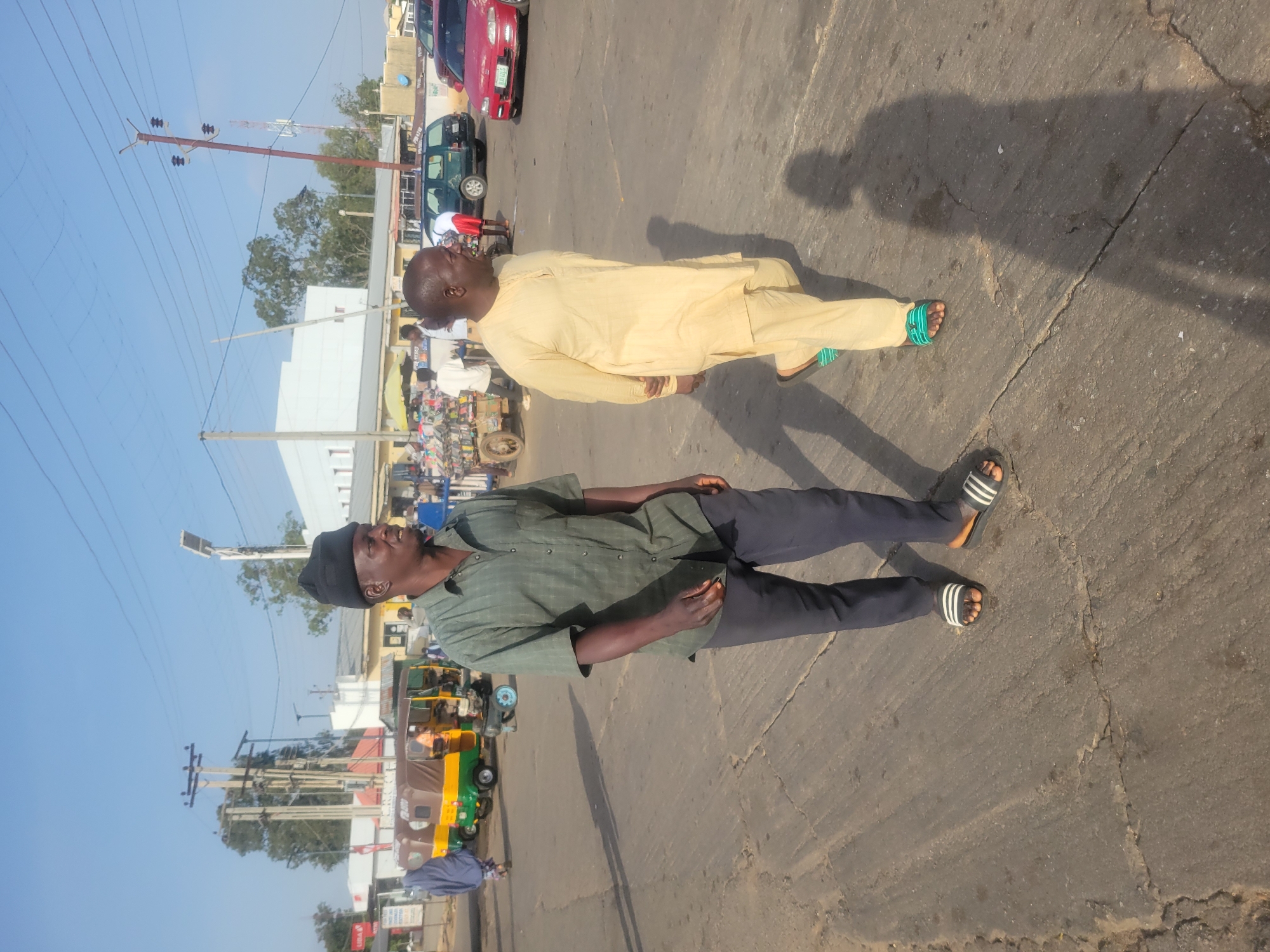 Two adult males are standing on a paved road. One wears a black beanie, a dark green button-up shirt, dark pants, and black and white sandals. The other wears a light yellow traditional outfit and light blue and white sandals. The road appears to be cracked. In the background, there are cars, buildings, power lines, and a three-wheeled vehicle called a "keke" or "tricycle taxi." The scene is likely in Jos, Nigeria, based on the presence of the keke, a common form of transport in that city. The photo was taken during the day, as shadows are visible.