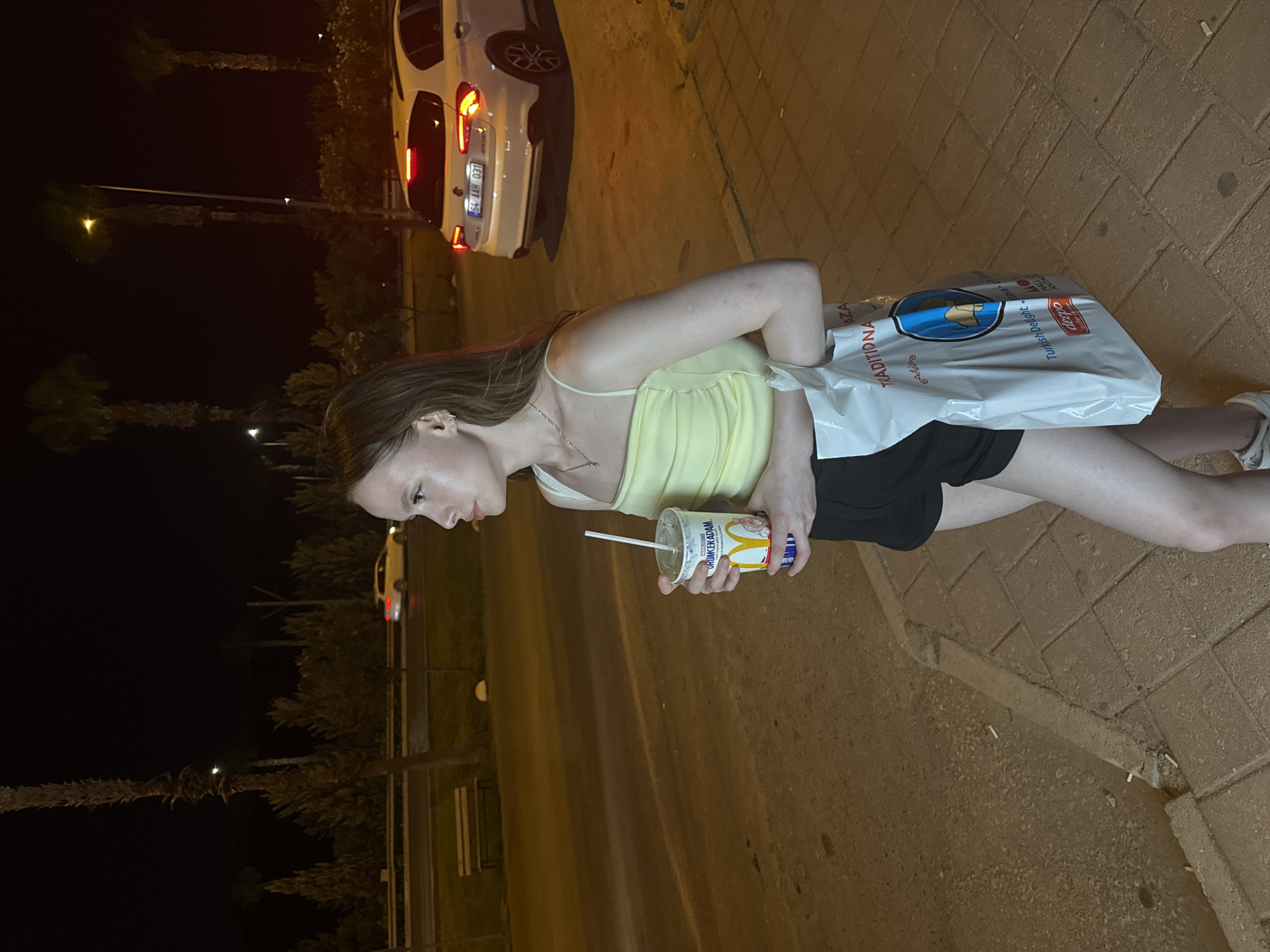 A young woman with light brown hair stands in the Kepez district of Antalya, Turkey, at night. She is holding a McDonald's beverage cup with a straw in her right hand and a white shopping bag with text and logos in her left hand. She is wearing a pale yellow crop top and black shorts. The background shows a street scene with palm trees, cars, and streetlights, suggesting an urban environment. One car, a white hatchback, is parked on the right side of the road with its brake lights illuminated. The ground is paved with concrete.