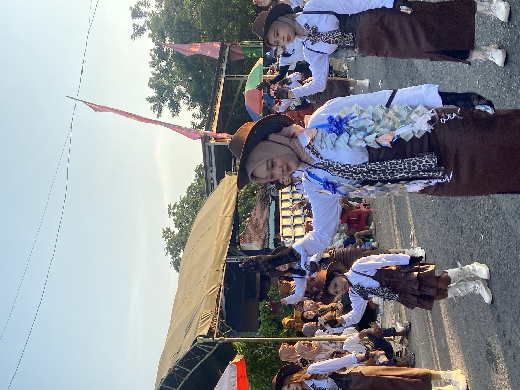 In the city of Margoyoso, Indonesia, a group of young women are participating in a parade. They are dressed in a coordinated outfit, consisting of a white long-sleeved shirt, brown skirts, and cowboy hats. Some are also wearing black gloves and white boots. Several of the women have money draped around their necks, which appears to be a form of decoration or performance. The parade is taking place on a paved road, with spectators gathered on the side. In the background, there are tents and trees, suggesting an outdoor event. The sky is clear and blue, with the sun shining brightly.