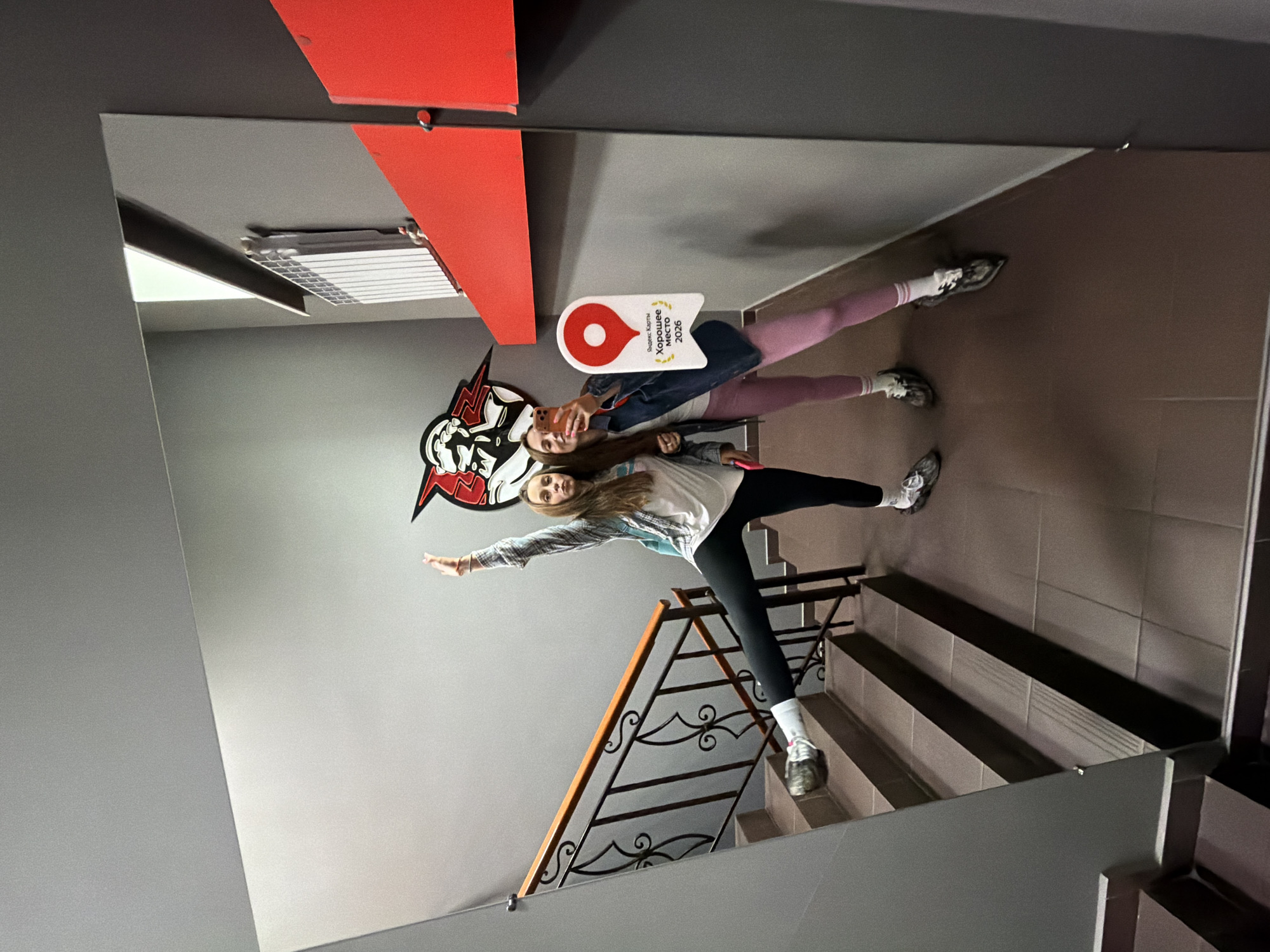 Two young women are posing for a selfie in a mirrored hallway, possibly in Birobidzhan, Russia, given the presence of a "Yandex Maps" sticker on the mirror. The woman on the left is standing on a stair, with her right leg extended forward and her left arm raised. She is wearing a plaid shirt over a white t-shirt, black leggings, and sneakers. The woman on the right is holding a phone, taking the selfie. She is wearing a denim jacket over a white t-shirt, pink leggings, and sneakers. They are both reflecting in a large mirror that covers a significant portion of the wall. In the background, a staircase with a dark metal railing is visible, along with a section of gray wall adorned with a graphic featuring a helmeted figure with red lightning bolts. Further to the right, a white radiator is mounted on the wall, and a red panel runs along the side. The floor is tiled in brown.