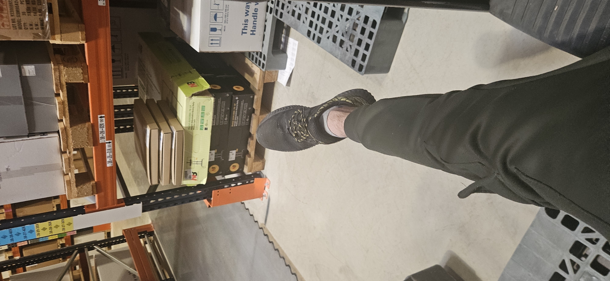 The image shows a person's leg and foot, clad in a black shoe and black pants, standing in a warehouse or storage area. Pallet racks filled with boxes and equipment are visible to the left. Some boxes have labels with text including "Handle with care" and "Mobile Cat". Labels on the orange pallet rack bars display numbers like "38-26-01X" and "38-26-02X" and other numbers with - followed by digits.
There is no mention or indication of the city of Plovdiv, Bulgaria.