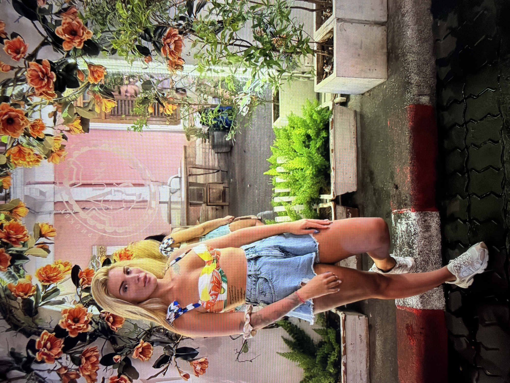 The image, correctly oriented, depicts a fair-skinned woman with long blonde hair posing outdoors, looking directly at the camera. She is wearing a colorful, floral-patterned bandeau-style crop top and light-wash denim shorts, complemented by white athletic shoes. Tattoos are visible on her arms and torso, and a light-colored scrunchie is on her left wrist.
The woman is positioned near a red and white striped curb, with a dark grey, patterned paving surface underfoot that appears wet. Artificial orange and yellow flowers on green vines extensively frame the left side of the shot. Various green plants and foliage are visible in white square planters to her right and in the background, suggesting a lush, possibly tropical, environment.
In the background, a light pink building or wall is present. Faint, light-colored text, partially obscured and blurry, appears on this pink surface, possibly spelling out "PHUKET" or a similar location name. Further back, wooden outdoor furniture, including a table and chairs, and a wooden balcony railing are discernible. The setting suggests a street, sidewalk, or an outdoor dining area, likely in a warm climate. The lighting indicates daytime, possibly overcast or early morning/late afternoon. The activity is a posed photograph.