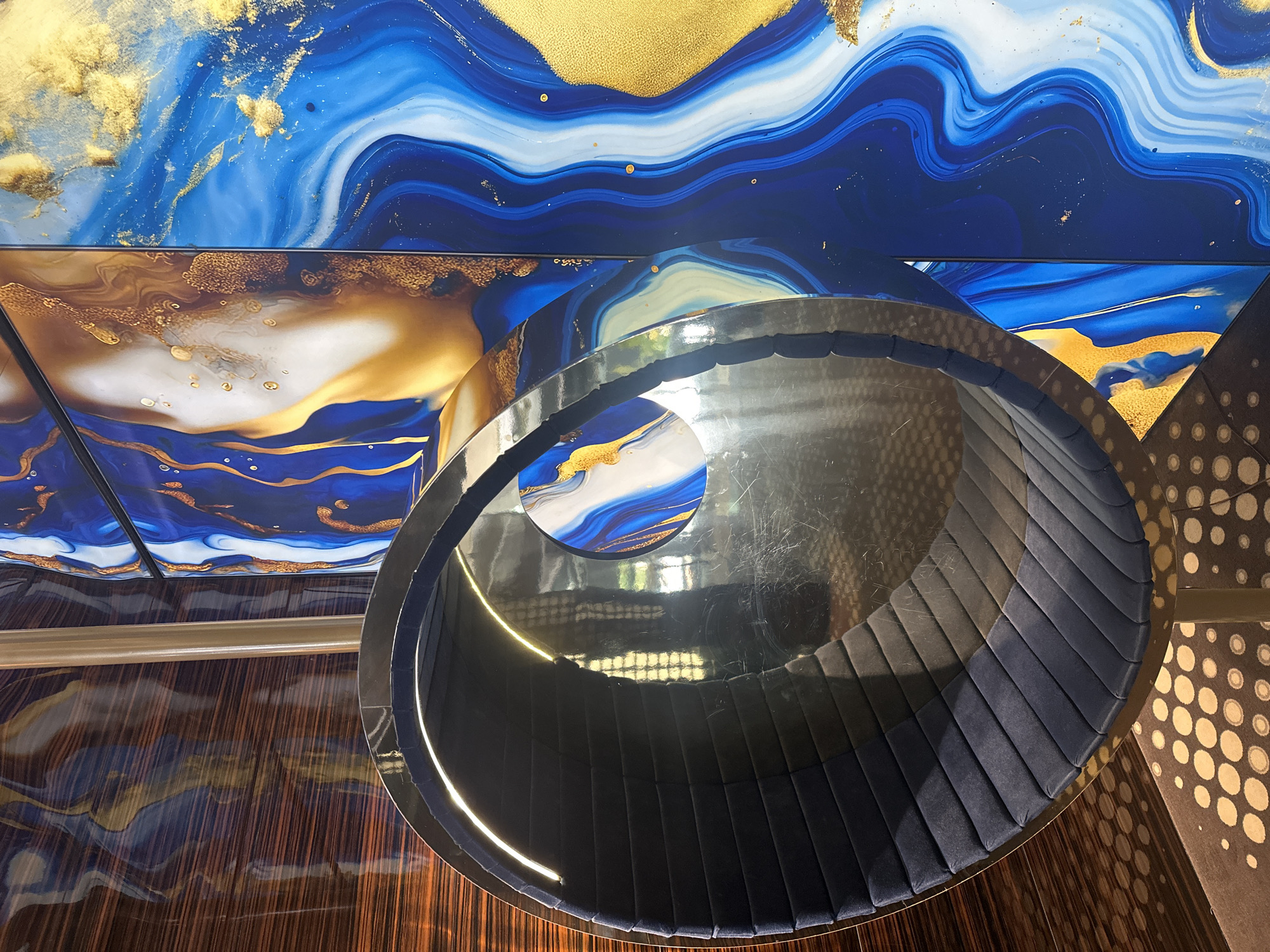 The image shows a close-up of a contemporary interior design. In the foreground, a dark blue upholstered circular chair with a metallic rim and interior lighting is positioned. The chair's interior is reflective. Behind the chair, a vertical, metallic rod extends upwards. To the right of the rod, a large wall mural with abstract patterns of deep blue, white, and gold is visible. The mural's surface has a glossy finish, with swirling lines and textured gold accents. To the left of the mural and partially obscured by the chair is a wall with a wood-like finish featuring horizontal striations in shades of brown and orange. The floor beneath the chair has a speckled pattern in beige and brown. The overall impression is of a modern, luxurious, and artistic space.