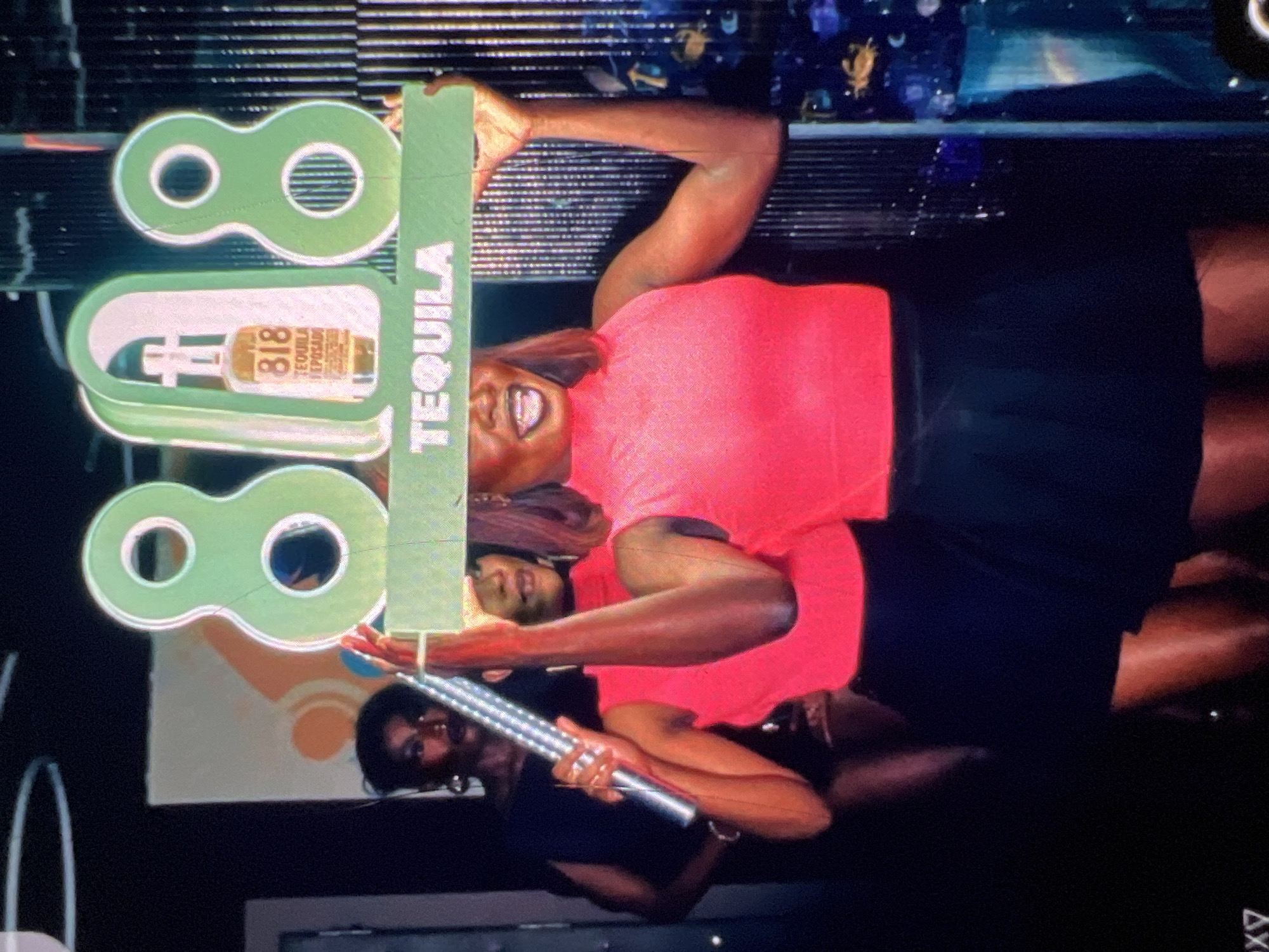 In Lagos, Nigeria, at a nighttime event, two Black women are the focus. The woman in the foreground, wearing a bright pink sleeveless turtleneck and a black skirt, holds up a large, illuminated sign that reads "818 TEQUILA." The sign features the number "8" on either side, with a stylized arch in the center framing a bottle of 818 Tequila. Behind her, another Black woman, wearing a black off-the-shoulder top, stands holding a silver, cylindrical object, possibly a microphone or a prop. The background is dimly lit, with vertical textured panels on the right and indistinct shapes and lighting elements suggesting a club or event venue.