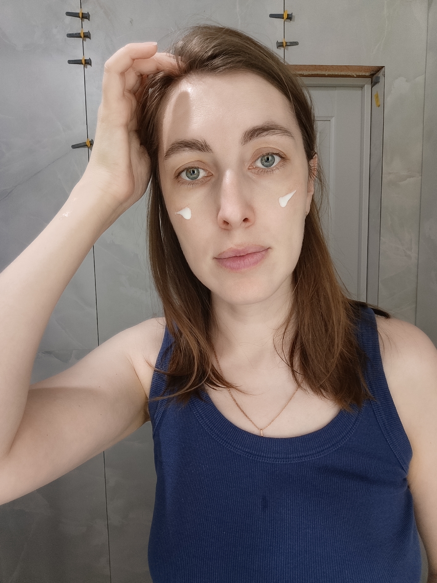 A woman with light skin, brown hair, and blue eyes is facing the camera. She has a dab of white cream on each cheek near her eyes. She is wearing a navy blue tank top and a gold necklace. Her left hand is raised to her hair. The background is a gray tiled wall. Construction materials (tile spacers) are affixed to the wall at the upper left. A partially visible door with a white panel is present in the upper right. There is no indication of location within Chaikovsky, Russia.