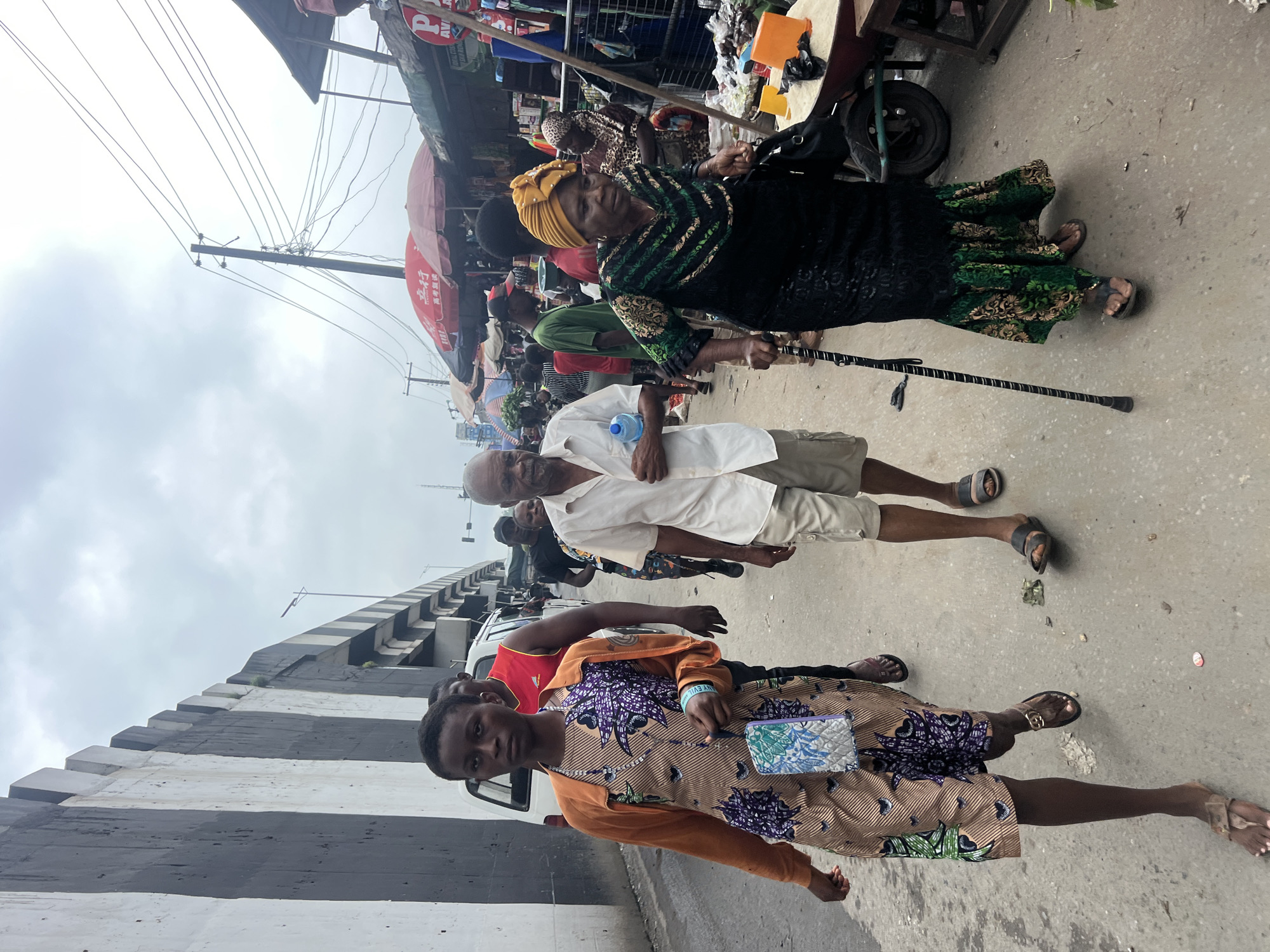 In Nkpor, Nigeria, a busy market street bustles with activity under an overcast sky. A concrete overpass looms on the left, its black and white stripes creating a stark contrast against the muted sky. Pedestrians navigate the uneven pavement, with a woman in a patterned dress and an orange cardigan leading the foreground. She clutches a small, patterned purse. Behind her, an older man in a white shirt and beige shorts walks with a cane, a bottle of water in his hand. Further back, a woman in a black dress and a yellow headwrap also uses a cane, her gaze directed forward. Stalls with red umbrellas line the right side of the street, displaying goods and attracting shoppers. The overall scene depicts a vibrant street market, a typical urban environment in Nigeria, filled with people going about their daily lives.