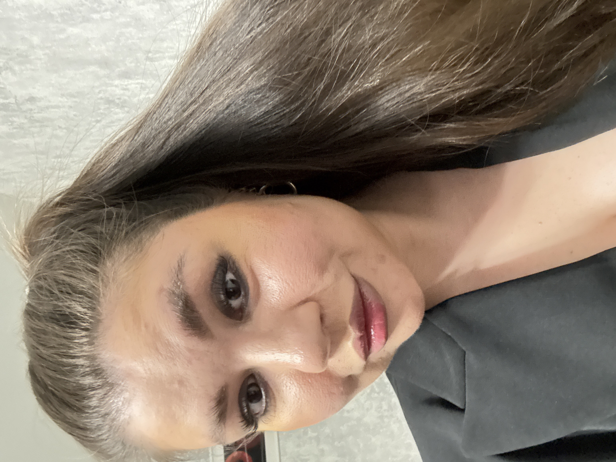 A woman is centered in the image, with the camera positioned at a slight upward angle to her face. She has brown hair, dark eyes, and fair skin. She is wearing a dark top. The background is a neutral grey color. The location is not identifiable from the image.