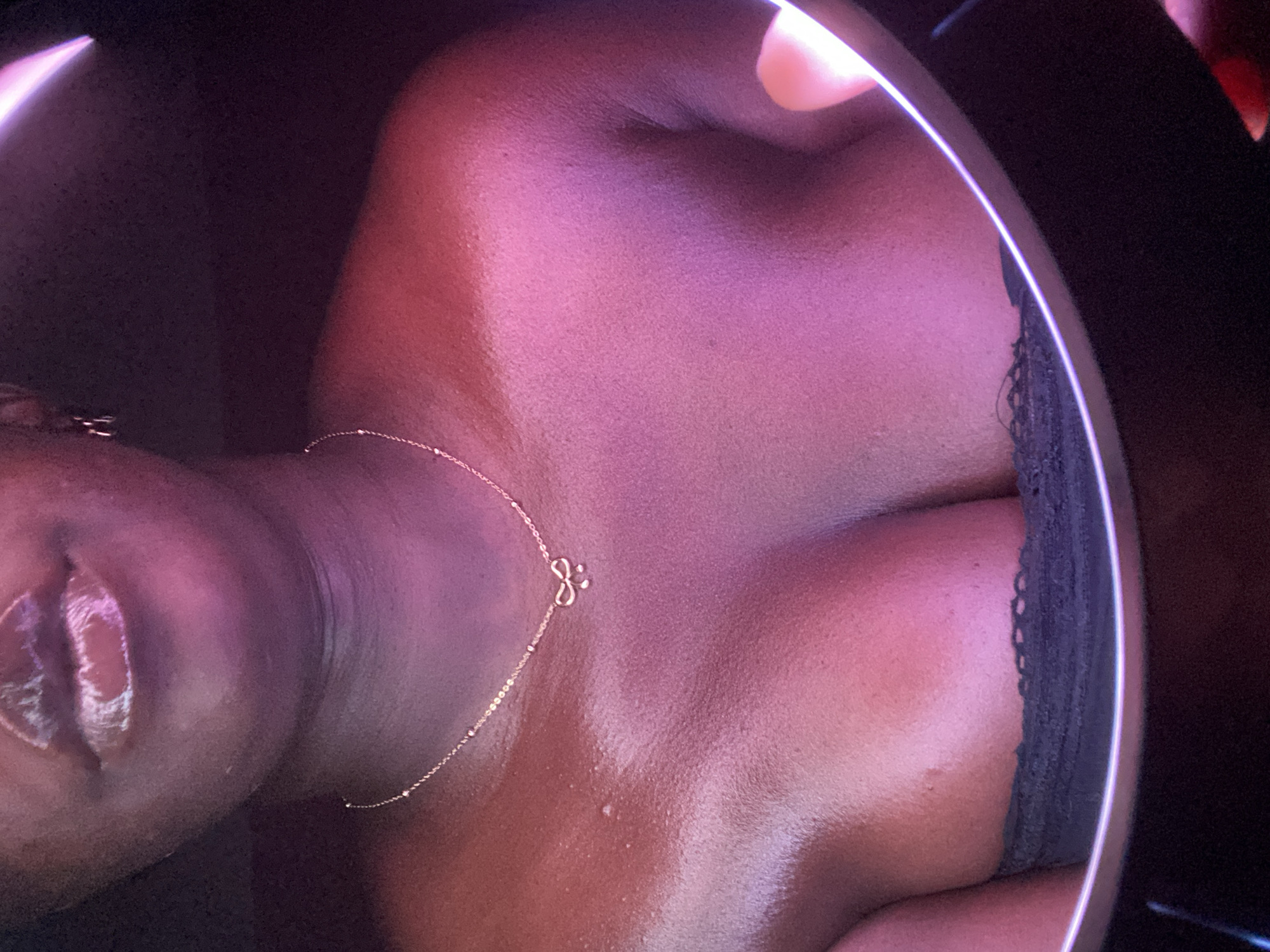 The image is a close-up, top-down shot of a person's upper torso, encompassing the chest, shoulders, neck, and part of the face, including the chin and glossy lips. The subject has a dark skin tone and is wearing a delicate, gold-colored chain necklace featuring a small, stylized 'S' pendant positioned on the sternum. A glimpse of a black lace garment, likely a bra or camisole, is visible on the right side of the frame.
The scene is dramatically illuminated by artificial light, casting vibrant pink and purple hues across the skin, giving it a dewy or moisturized appearance. The close-up perspective clearly shows the texture of the skin, including pores and some minor irregularities. A bright, circular light source, consistent with a ring light, is prominently reflected along the upper right edge of the frame, and another bright, angled light source is visible in the top left corner, contributing to the stylized and intimate atmosphere.
The activity depicted is likely posing for a self-portrait or a controlled photoshoot in an indoor setting. The lips are slightly parted and glossy, suggesting a sensual or alluring expression. Aside from the 'S' pendant, no other text is visible. While the given context places the location in Maigizo, Nigeria, there are no specific visual cues within the image itself to confirm this geographical detail. The lighting and composition create a mood of personal intimacy and aesthetic focus.