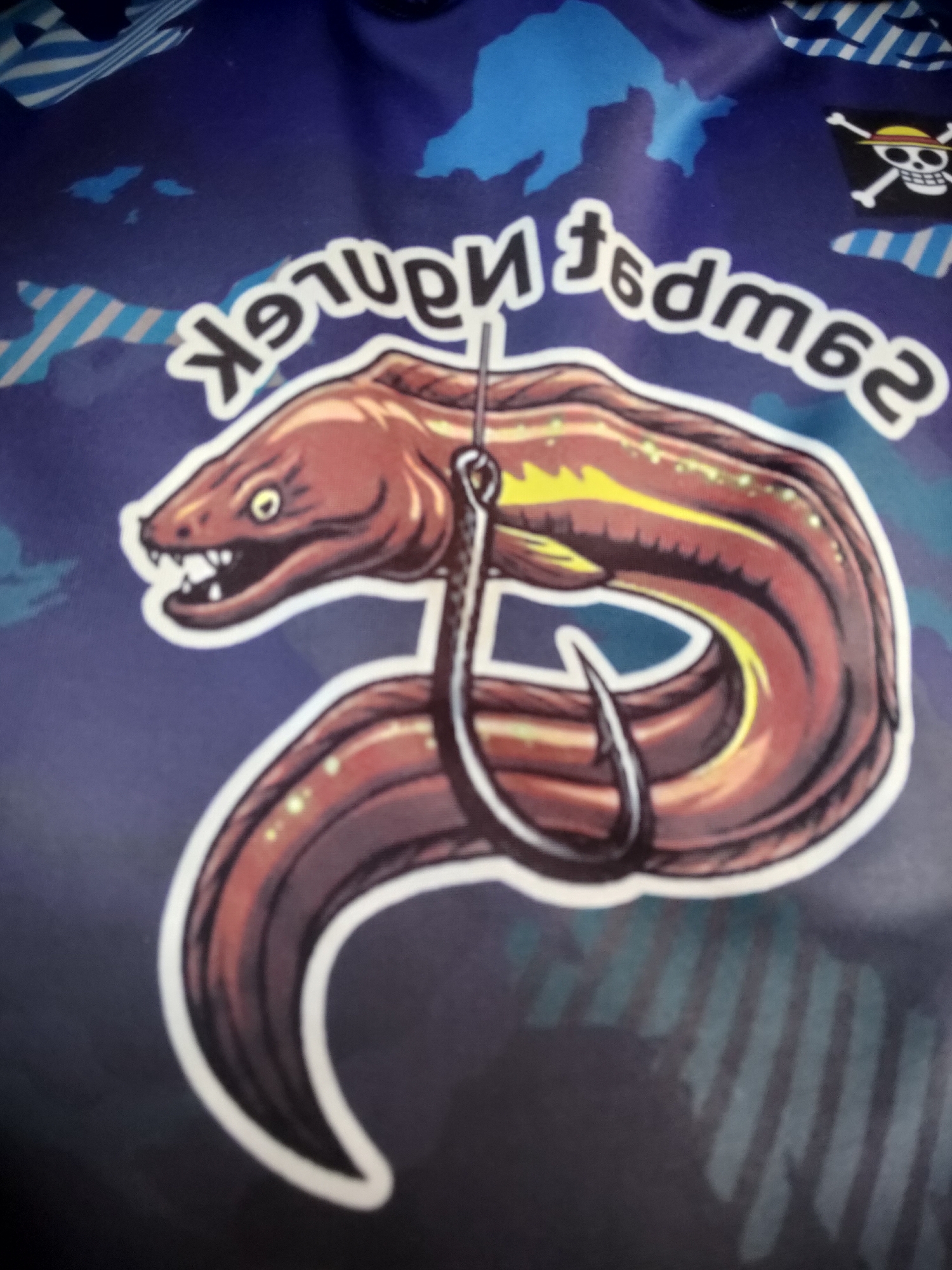 This is a close-up, eye-level, indoor photograph of a t-shirt with a graphic design. The design features a cartoonish moray eel with sharp teeth and yellow stripes, impaled on a fishing hook. The eel is predominantly brown with hints of yellow and green, and it is outlined in white. Behind the eel, the t-shirt displays a blue background with lighter blue shapes resembling landmasses. In the upper right corner of the graphic, there is a Jolly Roger flag, indicating a pirate theme. The text "SAWER NGEREDMES" is displayed in a white, stylized font arching above the eel. The photograph does not contain any elements that would place it in a specific city, such as Padang Dalom, Indonesia.