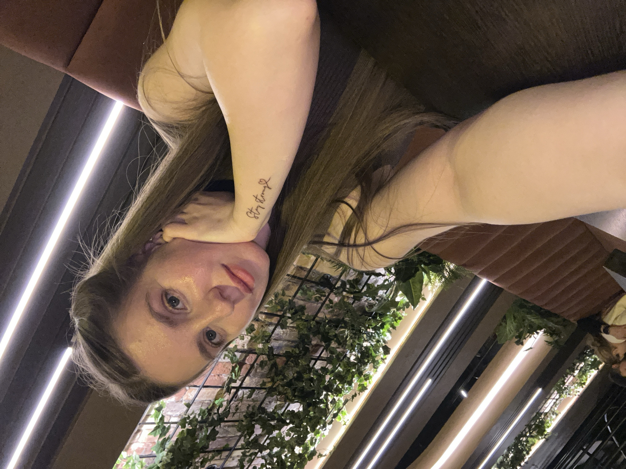 This media file presents a close-up portrait of a young woman in an indoor setting, likely a cafe or restaurant, located in Moscow, Russia. The original image is oriented horizontally, with the woman's head on the left side of the frame and her body extending to the right.
The woman has long, straight, dark brown hair that falls over her shoulders and back. She has fair skin and light-colored eyes, which are directed straight at the camera with a neutral, calm expression. She is resting her head on her left hand, with her left forearm visible in the foreground. On her left forearm, a small, black script tattoo reads "Stay Strong" followed by a tiny heart symbol. Her right shoulder and a portion of her upper arm are also visible, suggesting she might be wearing a light-colored top.
The background elements contribute to a modern and stylish ambiance. Behind her, a dark brown, possibly upholstered, bench or booth seating is visible. Further back, a decorative arrangement of green, ivy-like plants on a black trellis covers a section of a reddish-brown brick wall. The space is illuminated by several linear, bright white LED lights, which appear to be integrated into the ceiling or wall structures, creating a contemporary glow. Dark panels and structural elements are also visible, contrasting with the bright lights and greenery.
The scene captures a relaxed moment, possibly a selfie or a casual photo taken in an interior space during the evening or night, given the reliance on artificial lighting.