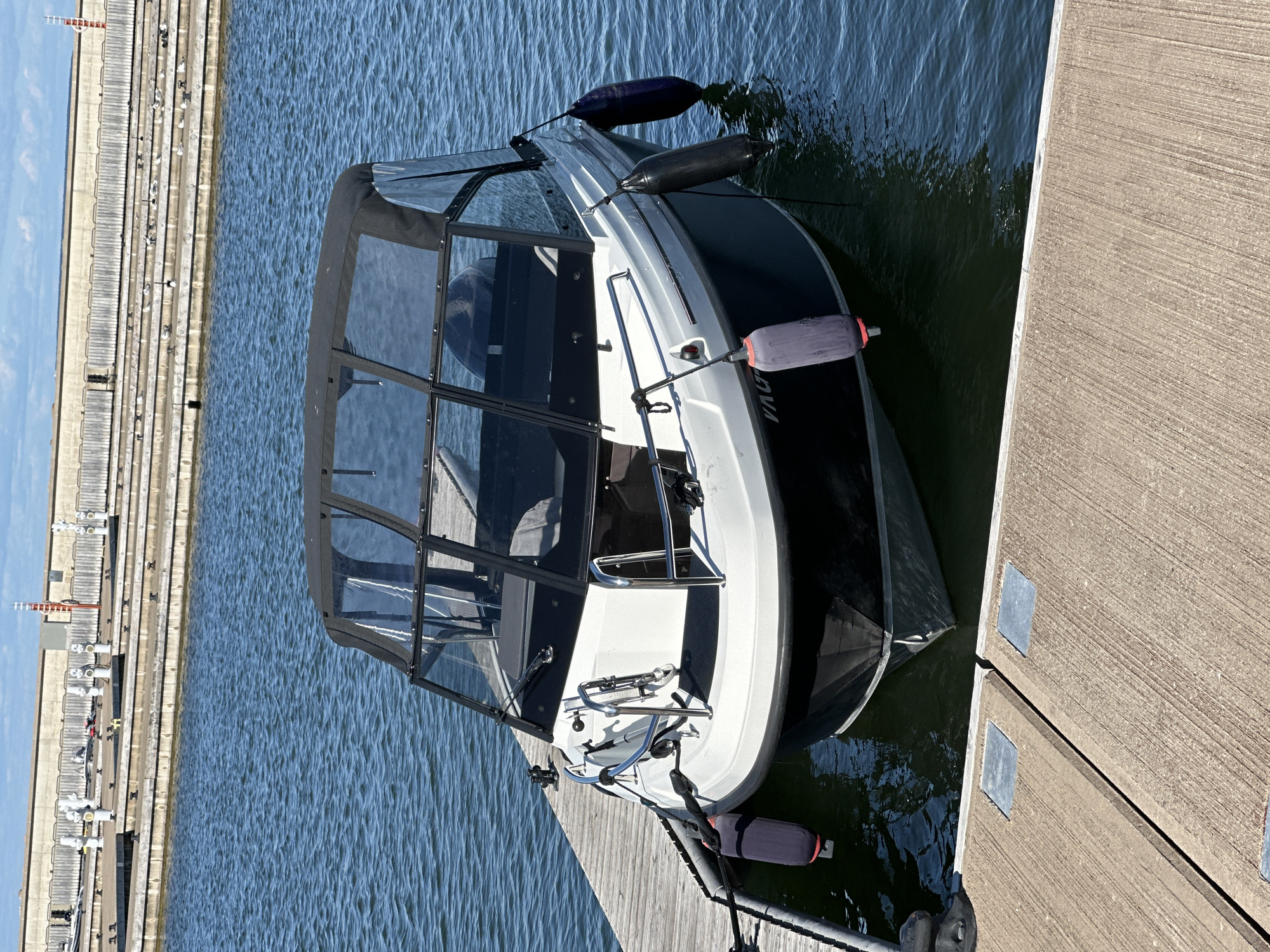 A white and dark blue motorboat with a black canvas cover is docked at a wooden pier on a clear, sunny day. The boat is secured with ropes and has several dark blue and black fenders attached to its sides. The water is calm and blue. In the background, a concrete seawall with tiered seating and various structures, possibly part of a marina or port, is visible. There are no people visible in the image. The boat appears to be in good condition and ready for use. The scene is likely in a harbor or marina area in Tallinn, Estonia.