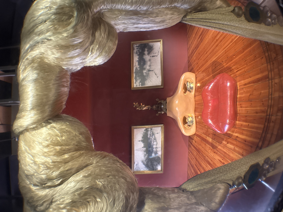 The image captures an interior space, specifically an art installation or a uniquely designed room, viewed from a slightly elevated perspective. The photograph itself is rotated 90 degrees counter-clockwise, making the floor appear as a right-hand wall and the background wall as a top wall.
When viewed in its intended orientation, the scene features a surreal and elaborate aesthetic. To the left and curving around the top, massive, intertwined strands of golden-blonde, lustrous hair form a prominent part of the decor, resembling an elaborate wig or a canopy. These strands are thick, textured, and reflect light, suggesting a synthetic material like nylon or silk.
The main wall in the background is a solid, dark red or maroon color. Hanging on this wall are two rectangular, gold-framed pictures. The images within the frames are black and white or sepia-toned and appear somewhat abstract or historical, possibly depicting architectural structures or landscapes with a grainy texture. Between these two framed pictures, centrally placed on the wall, is a small, intricate, brass-colored decorative object, possibly a figurine or an ornate sconce, with delicate details.
Below the wall, extending across what would be the floor, is a distinctive wooden surface. This floor is made of reddish-brown wood planks arranged in a radial pattern, fanning out from a central point, creating a sunburst effect. On this wooden floor, two large, sculptural elements are prominently displayed. To the left is a flesh-toned, oversized representation of a human nose, with golden, possibly tooth-like, details visible within the nostrils. To the right of the nose is a large, glossy, red object shaped like a pair of sensuous lips, appearing inflated or cushion-like, similar to an inflatable toy or a piece of pop art furniture.
On the far right side of the image, running diagonally across what would be the lower part of the scene, is a section of draped fabric. This fabric is a muted olive or khaki green, with decorative gold and possibly green jeweled embellishments or brooches attached, suggesting a rich curtain or upholstered furnishing.
The overall impression is one of a highly stylized and whimsical, possibly surrealist, environment, reminiscent of the works of Salvador Dalí. The lighting is even, suggesting an indoor setting with controlled illumination, highlighting the various textures and forms within the installation. There are no people visible, and no discernible text.