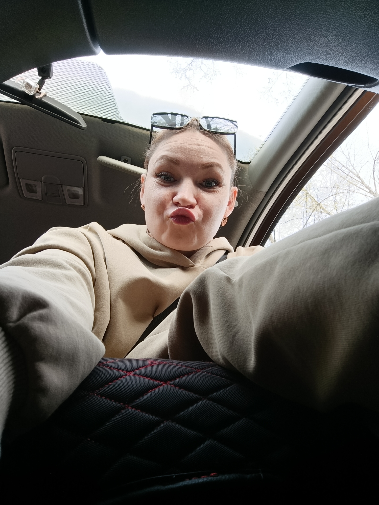 A woman takes a selfie inside a car. She is wearing a tan-colored hoodie and has sunglasses perched on top of her head. She is making a kissing face. The car interior has a black seat cover with red stitching in a diamond pattern. Through the car window, bare trees can be seen. Based on the metadata, this image was taken in the city of Chaikovsky, Russia.
