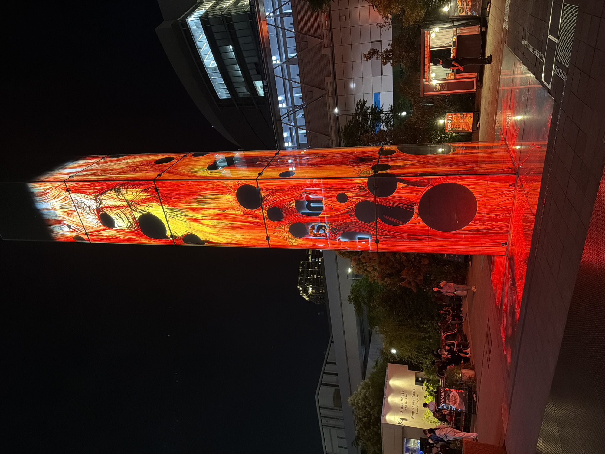 This is a night-time, eye-level, outdoor shot of a vibrant, illuminated, rectangular tower displaying abstract, fiery red and yellow patterns with black circles. The tower is the focal point, located in a plaza. To the left, a low-lying building with signage for "Glass House" and "Emptiness Table" is visible, with several people seated on benches in front of it. A "Ramen" sign is also present. Further to the right, a food truck or stand with a sign advertising "DONUT" is situated, with a person standing behind the counter. In the background, modern buildings with illuminated windows are visible. The ground is paved, and the tower casts a red reflection onto the wet-looking surface. The sky is dark, with a few faint stars. This scene is located in Koto City, Japan, specifically at the Toyosu Market's new urban development area, "TOYOSU PIT."
