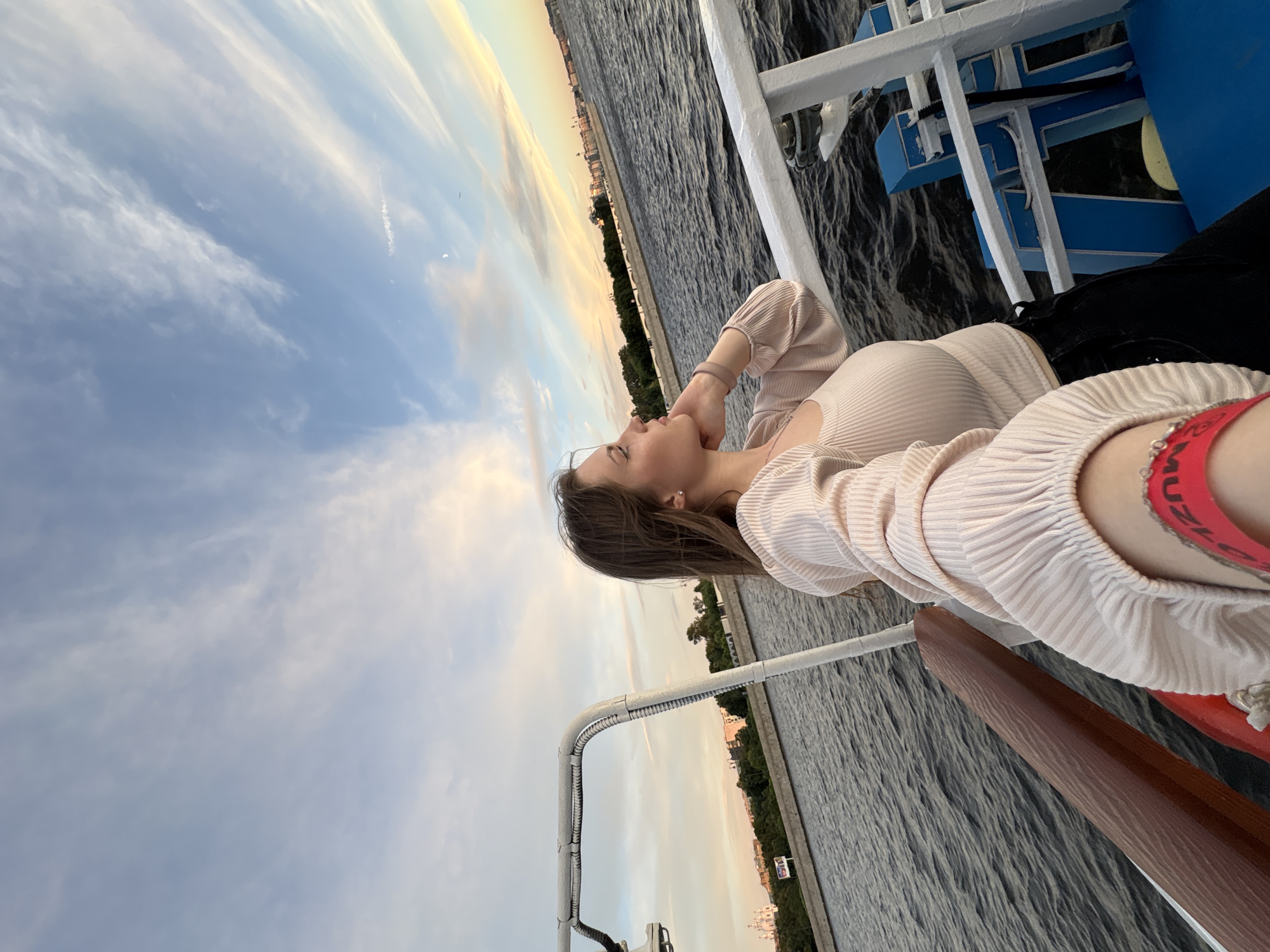 The image shows a young woman taking a selfie on a boat. She is wearing a light pink ribbed top and black pants. She is looking out at the water, with a city skyline in the background. The sky is a pale blue with wispy white clouds, and the sun appears to be setting, casting a warm glow over the scene. The water is dark and choppy, with small waves. The boat appears to be a tour boat, with white railings and blue seating. In the background, a city skyline is visible, with buildings and trees lining the waterfront. The architecture suggests it is likely the city of Saint Petersburg, Russia, possibly along the Neva River or one of its canals, given the presence of ornate buildings and a general sense of historical grandeur. The overall impression is one of a relaxed and scenic boat ride.