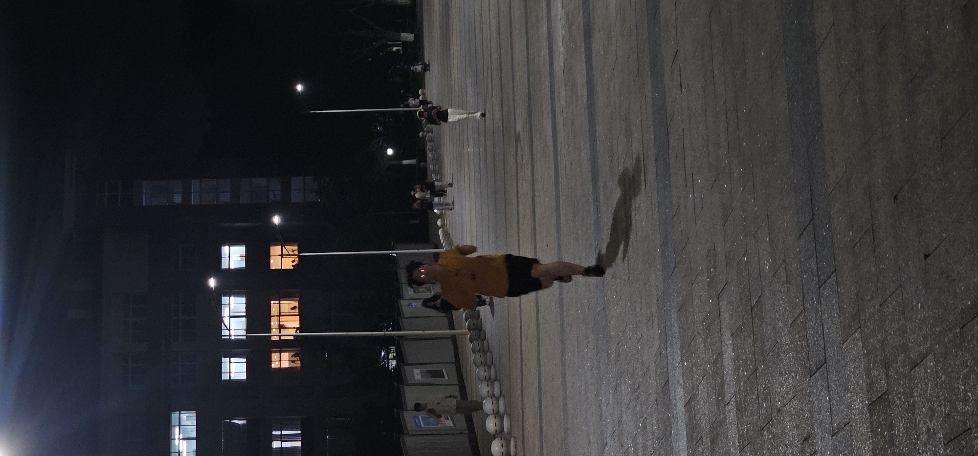 At night in Chongqing High-tech Zone, China, a person in a yellow shirt and black shorts stands on a paved area with a dark, building-like structure on the left. Windows in the building are lit. Streetlights are visible. There are other people in the background. The person casts a shadow on the pavement. The image has been rotated.