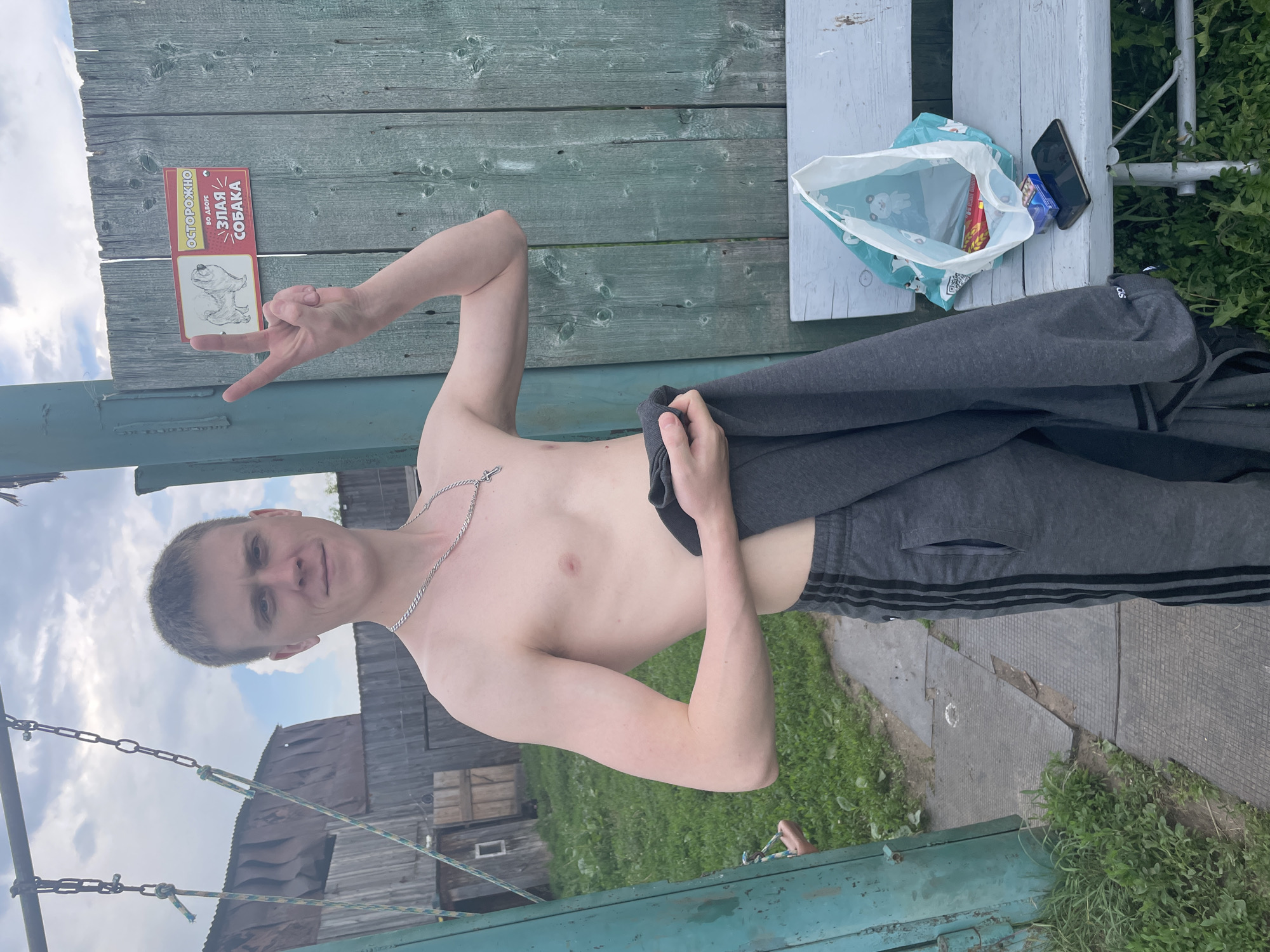 A young, shirtless Caucasian male with short, light brown hair and fair skin stands in front of a weathered, teal-painted wooden fence. He is wearing grey athletic pants with black stripes down the sides. He holds a dark grey garment in his left arm and extends his right arm, making a peace sign with two fingers. A sign on the fence reads "ОСТОРОЖНО ВО ДВОРЕ ЗЛАЯ СОБАКА" (Caution, beware of the fierce dog) with a drawing of a dog. To his left, a metal swing set with chains is visible, and behind him are wooden buildings and overgrown grass. To his right, a light blue wooden bench sits on the grass. On the bench are a plastic bag with items inside, a pack of cigarettes, and a black smartphone. The scene appears to be an outdoor, rural residential area, possibly a dacha or village in Nizhnii Bugrysh, Russia, given the context. The lighting suggests it is daytime, with a cloudy sky overhead.