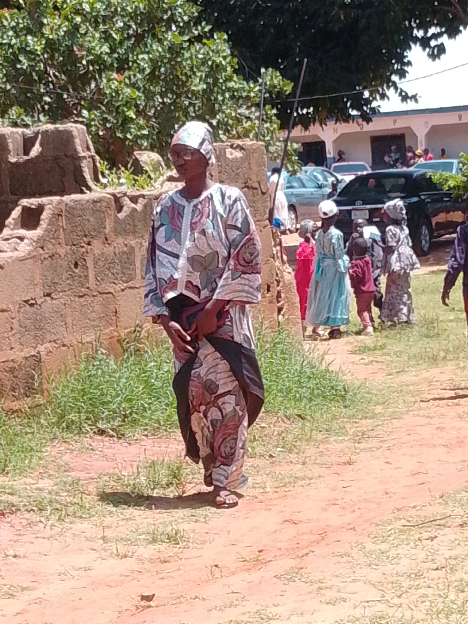 An outdoor, full-shot photograph captures a woman in the foreground walking on a dirt path in Tumfure, Nigeria. She is dressed in a traditional, patterned outfit with a matching headscarf and glasses, her hands are clasped together in front of her. Behind her, a group of children, also in traditional attire, are gathered near a building. The scene is illuminated by natural sunlight, casting shadows on the ground, and is framed by lush green vegetation and a brick wall. The background shows a building with cars parked in front, suggesting it might be a public space or an event. The overall ambiance of the photograph is that of a sunny day in a community setting.