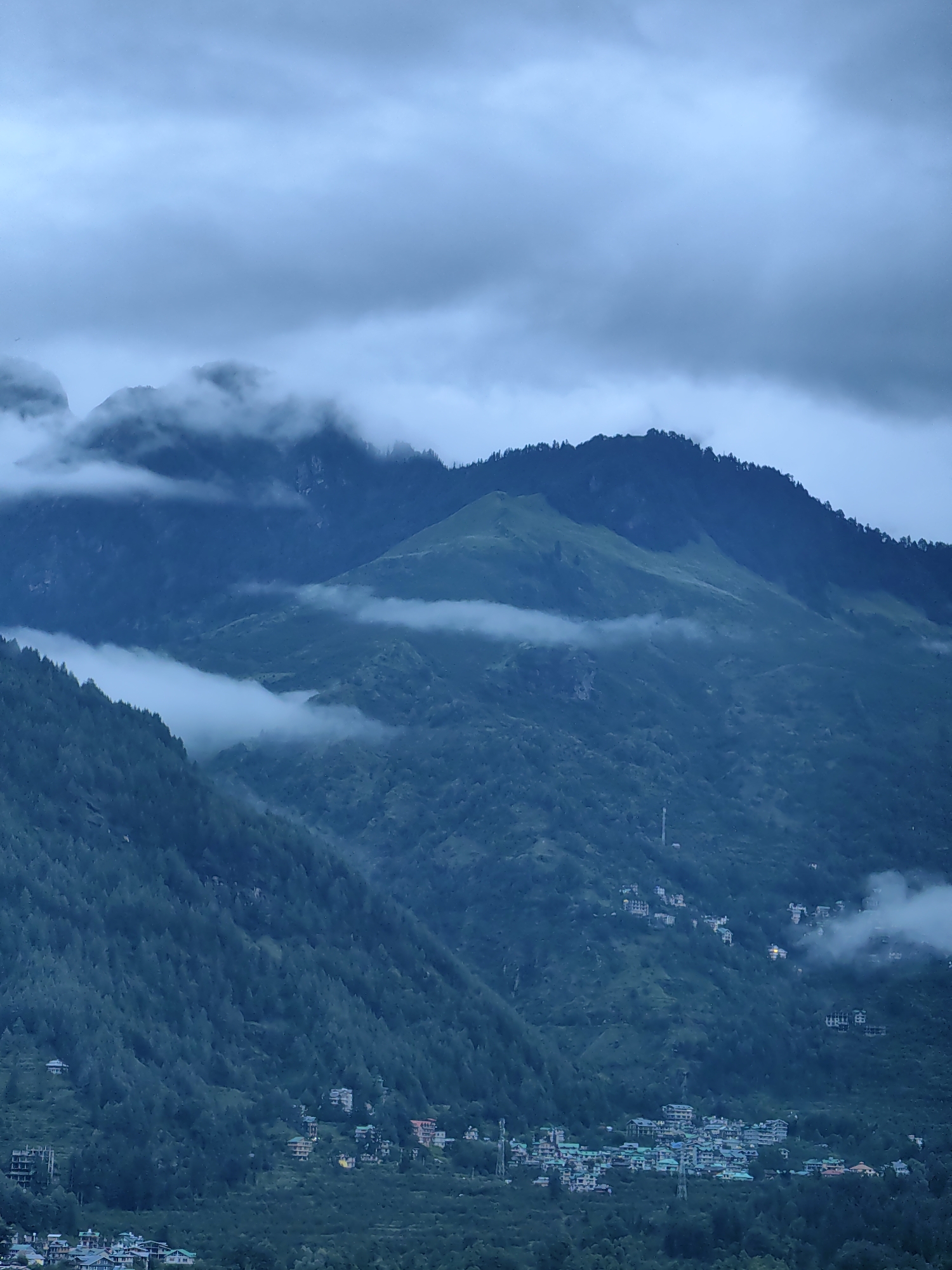 This image depicts a scenic view of the town of Vashisht, India, nestled amidst lush green mountains. The sky is overcast with thick clouds, casting a dim, moody atmosphere over the landscape. The foreground is dominated by dense coniferous forests that carpet the steep slopes of the mountains. Scattered amidst the trees and on the lower slopes are clusters of buildings, indicating the presence of the town. These structures appear to be mostly multi-story buildings with colorful roofs, suggesting a residential or tourist-oriented area. Some buildings are more visible than others, peeking through the foliage. Wisps of fog or low-lying clouds cling to the mountainsides, adding to the ethereal quality of the scene. The overall impression is one of a remote, mountainous settlement shrouded in mist and natural beauty. There are no visible people or animals in the image.
