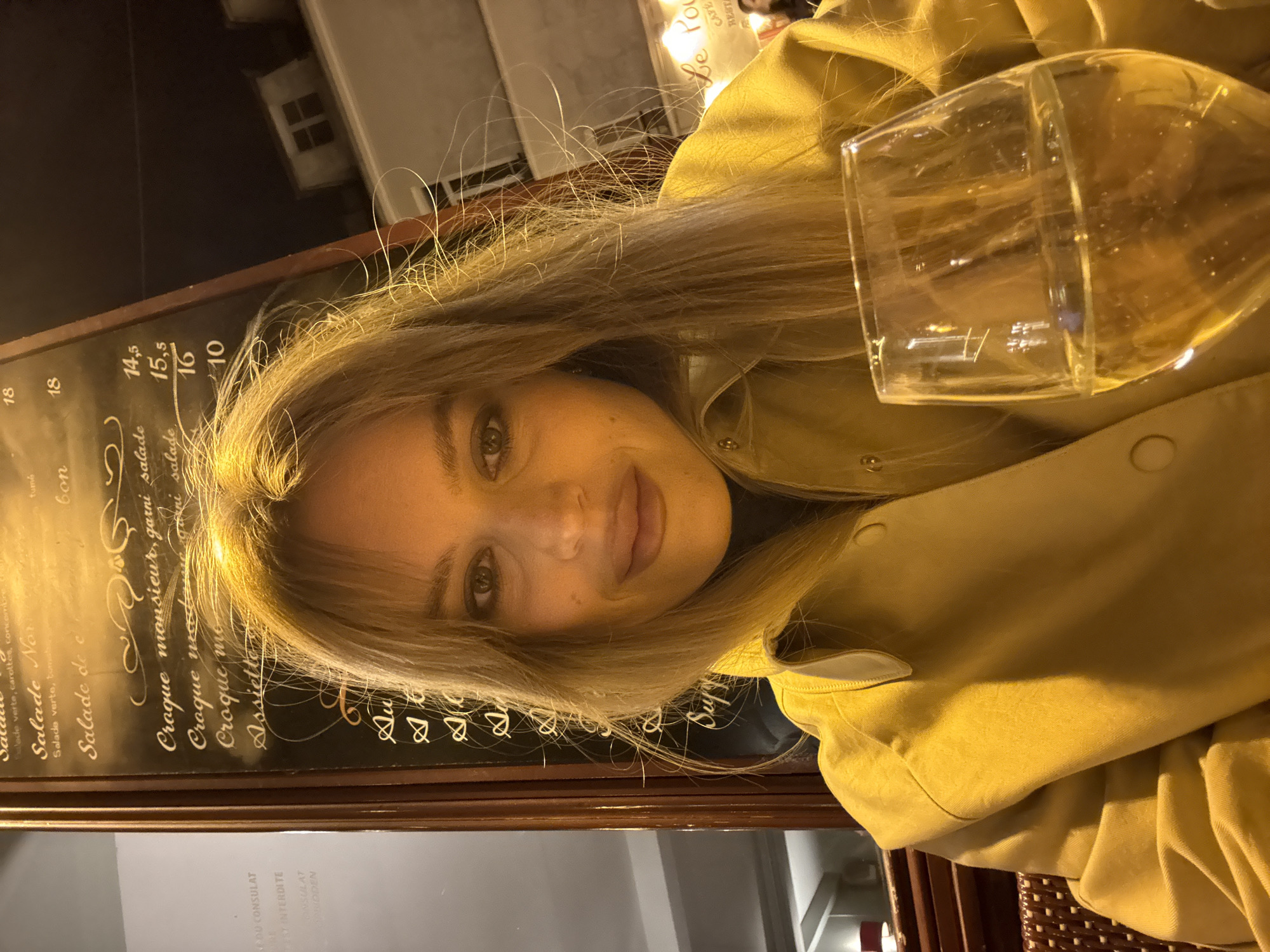 The photograph captures a woman at an outdoor café, potentially in Paris, France, given the appearance of a French menu board to her left, listing items such as "Salade de" and "Croque monsieur, garni salade." She is holding a glass of a clear liquid, likely wine. She is dressed in a light-colored jacket, and her light brown/blonde hair is visible around her face. In the background, a building with a window can be seen. The illumination suggests it is evening or nighttime. The menu board lists prices in euros for various items, as well as the name of the establishment, "Le Pou." The style of the menu board and the surrounding architectural details are consistent with a typical Parisian café setting.