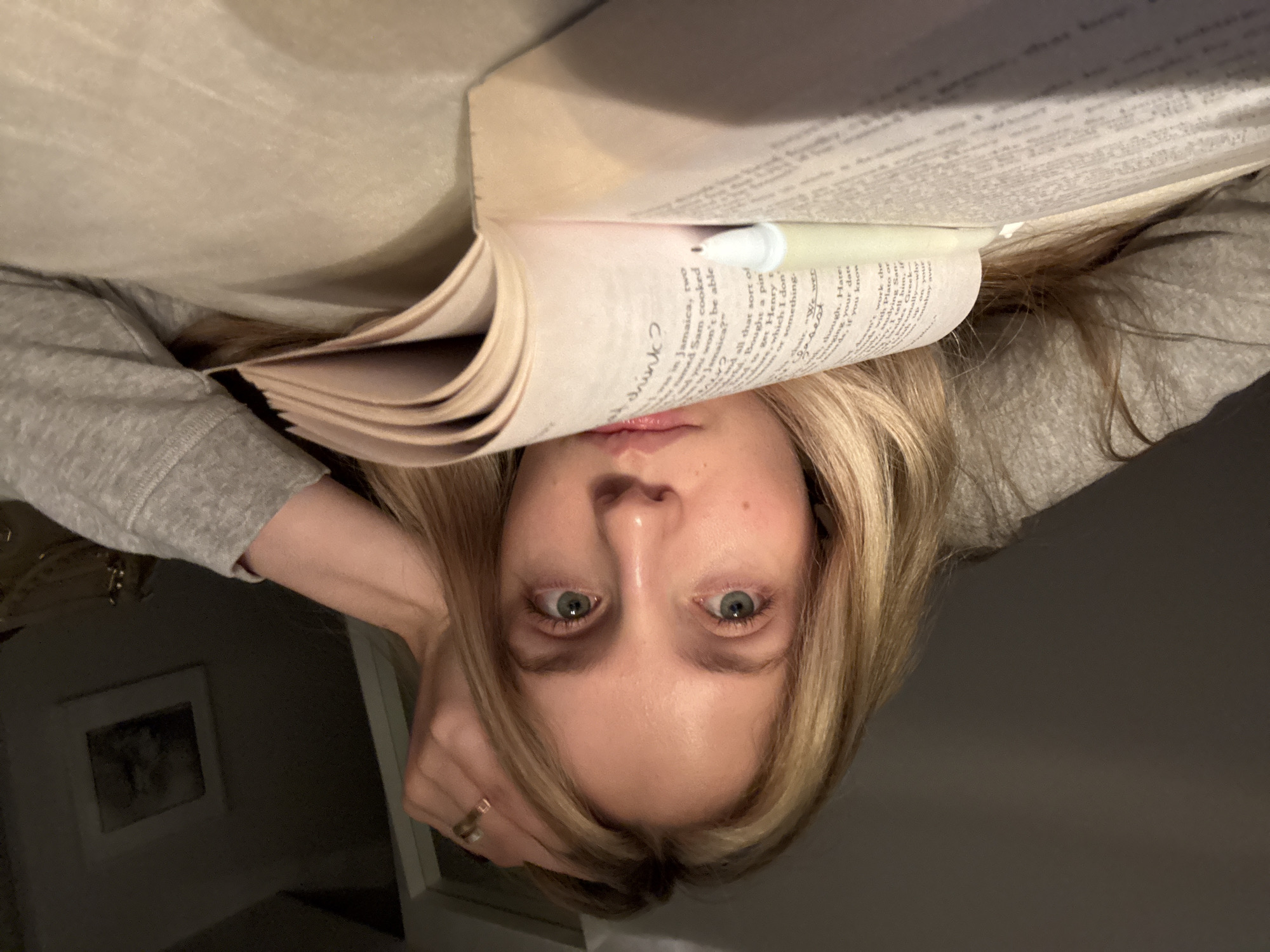 The image presents an upside-down view of a woman lying down and reading a book. The book is open, positioned above her face with a pen marking a page. The text on the page is partially legible, discussing a setting in Jamaica and mentioning names like Sam. The woman's face is visible, her eyes gazing forward. She is wearing a gray long-sleeved shirt, and a ring is visible on her hand. In the background, a framed picture hangs on the wall. The location, Mexico City, Mexico, is not apparent from the visual information provided in the image.