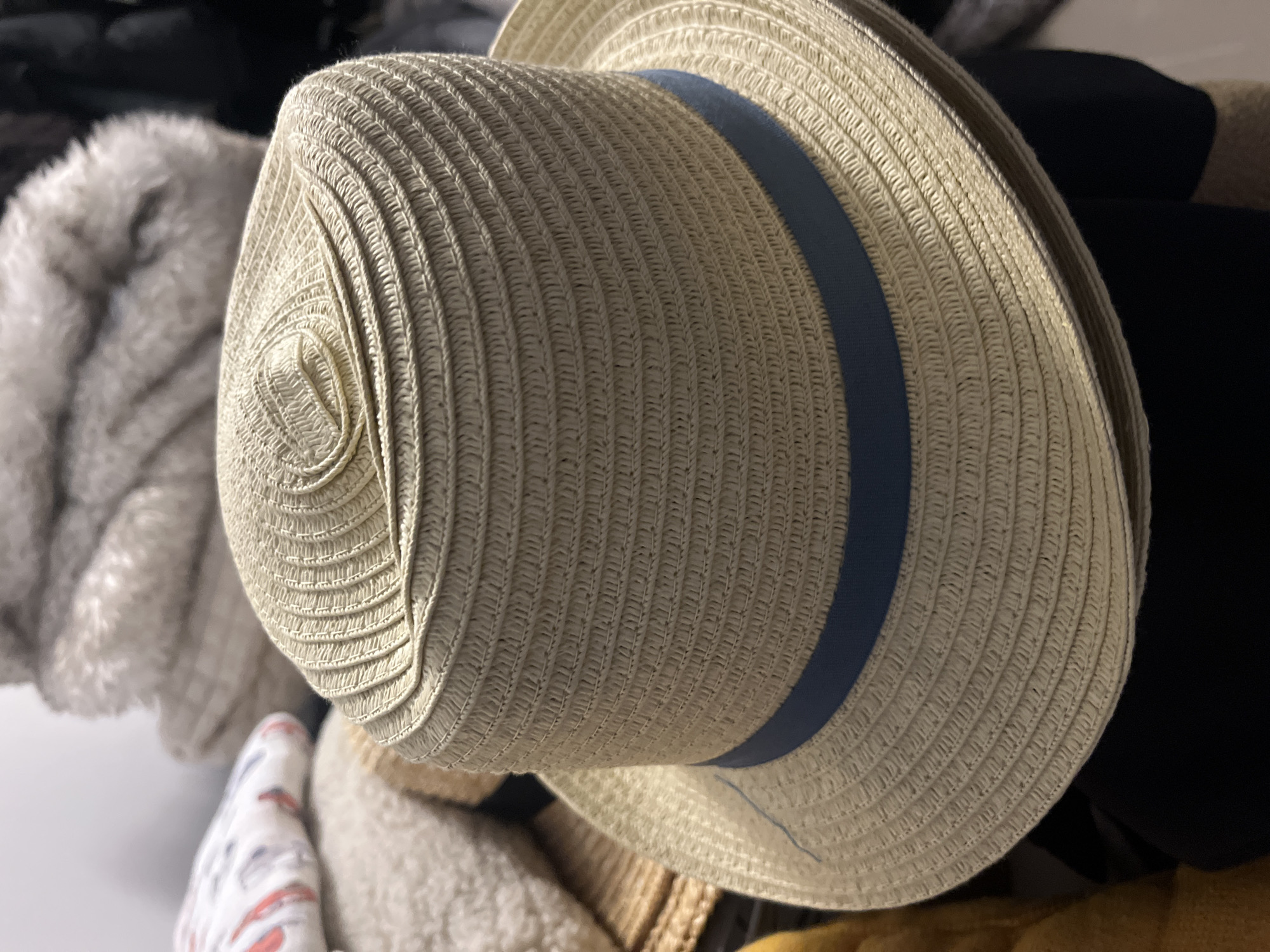 A close-up shot focuses on a woven straw hat with a blue band. The hat is positioned in the foreground, angled slightly to the left, with its crown and brim clearly visible. The weaving pattern of the straw is intricate, with concentric circles forming the crown. In the background, out of focus, are piles of soft, textured materials, possibly clothing or blankets, in neutral tones of white and beige. The lighting appears to be soft and even, highlighting the texture of the hat. There is no indication of any specific location in the city of Onokivtsi, Ukraine.