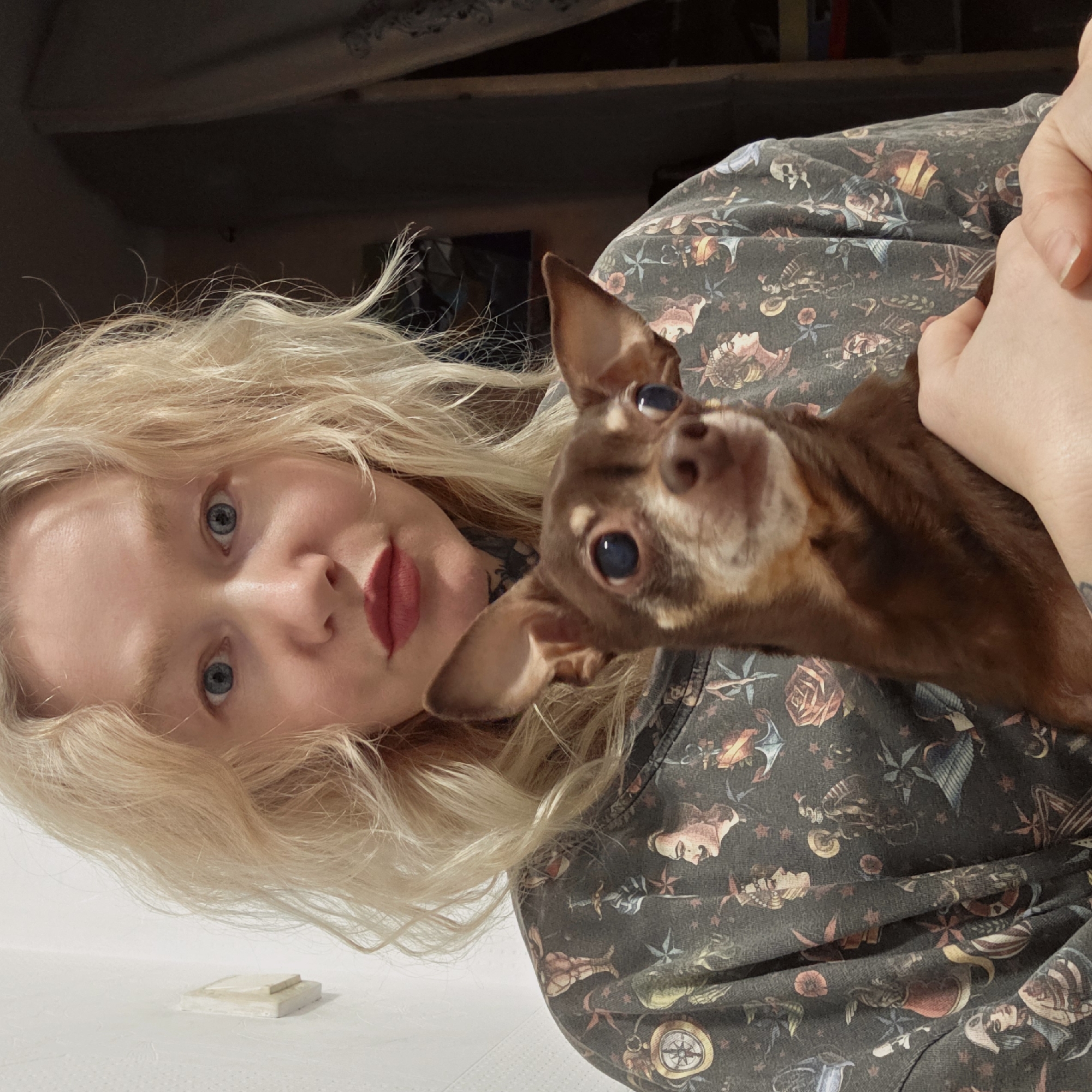 A person with fair skin, blonde hair, and red lipstick is holding a brown Chihuahua-like dog. The person's shirt has a pattern of vintage tattoo-inspired imagery, including roses, compasses, and human faces. The background is a partially visible room with light walls. The image shows no details to suggest it was taken in Saint Petersburg, Russia.
