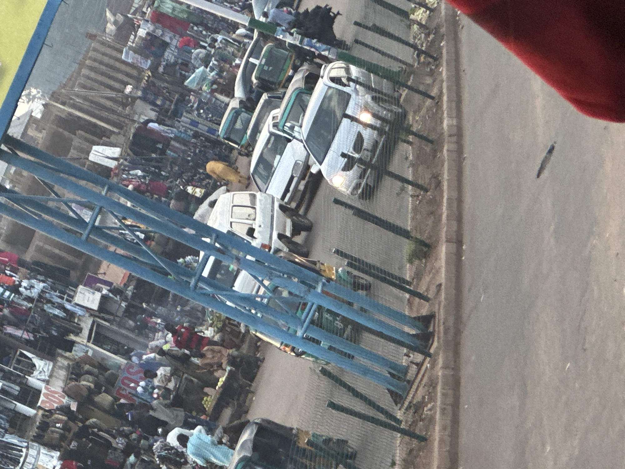 The image captures a busy street scene in Bukuru, Nigeria, likely a market area. In the foreground, a blue metal structure, possibly part of a canopy or scaffolding, dominates the left side, partially obscuring the view. Beyond this structure, several vehicles are visible on the paved road.
To the left, a black auto-rickshaw is parked. Further ahead, a green and yellow auto-rickshaw is in motion. A white van is parked to the left of a white sedan which is in the foreground and is silver in color. Behind the white sedan, a gray sedan is visible. Other vehicles, including more auto-rickshaws and cars, are seen further down the road.
The background reveals a bustling marketplace with numerous stalls and vendors. Clothing and various goods are displayed, indicating commercial activity. Several individuals are present, some appear to be shoppers or vendors. A person in a yellow garment is walking away from the camera. The overall impression is one of lively urban commerce.