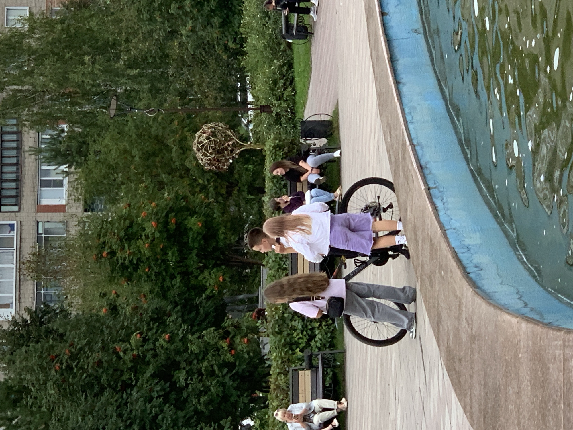 In a park in Prokopyevsk, Russia, three young people are gathered around a bicycle near a fountain. A boy with short brown hair stands to the right, looking away from the camera and holding what appears to be a small object. To his left, a girl with long brown hair, wearing a pink shirt and grey pants, stands facing forward. Beside her, a girl with blonde hair and wearing a white top and purple shorts stands facing the boy. The bicycle, a mountain bike with a black frame, is positioned between them. In the background, an older woman sits on a park bench, looking at her phone. Other individuals are visible on benches further in the distance, and trees and a building are present in the background. The fountain has a concrete edge and the water is a murky green color with white foam.